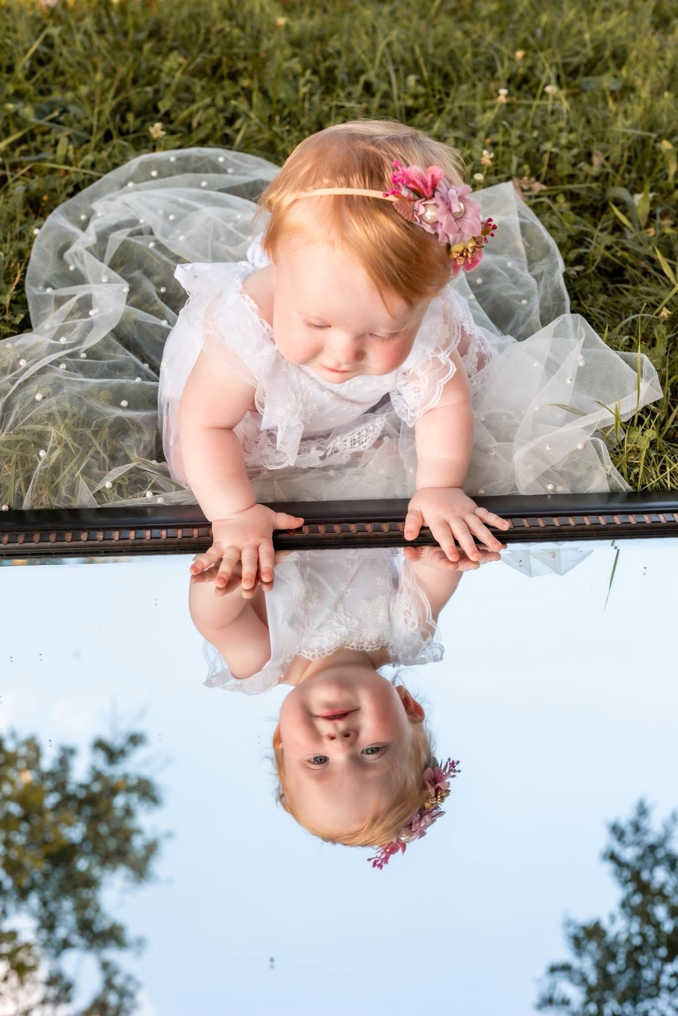 Creative mirror reflection photo of baby in white dress on grass.