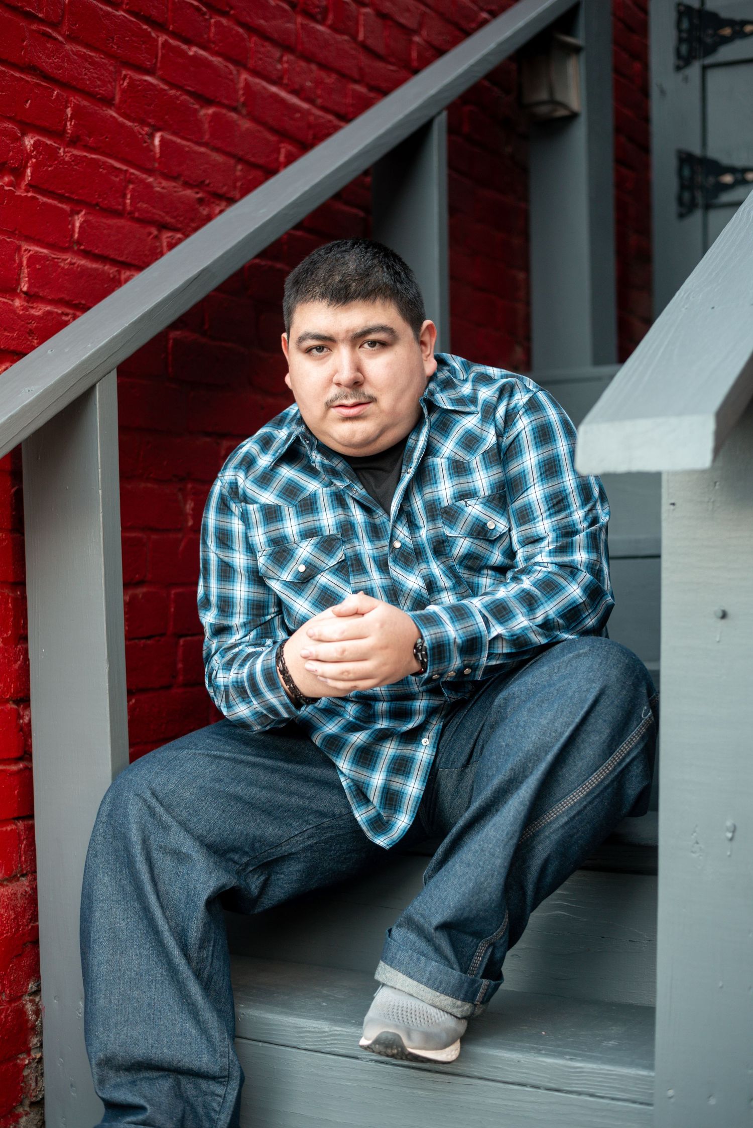 Person wearing blue plaid shirt and jeans sits casually on gray steps against red brick wall.
