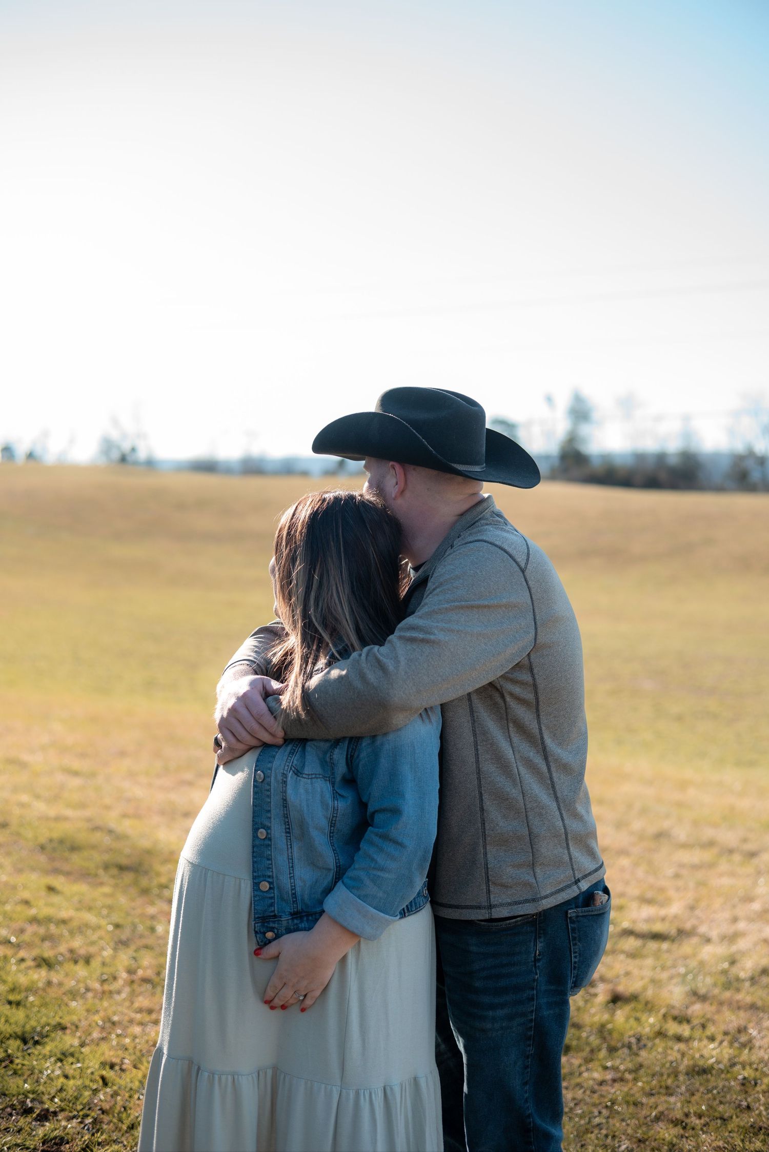 A couple embraces in a sunny field wearing casual western attire during a maternity photoshoot.