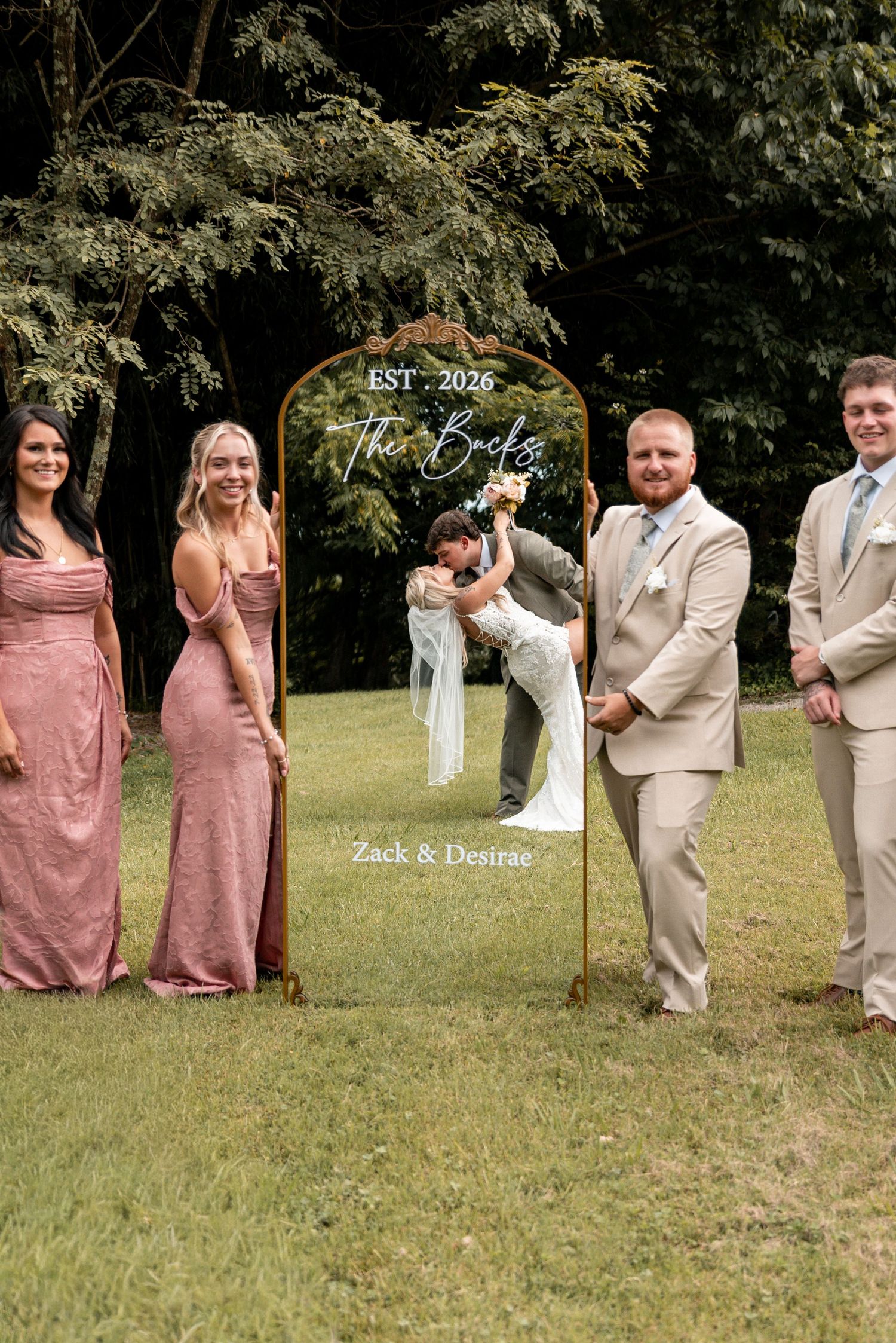 Wedding party poses outdoors with a decorative mirror sign reading The Bucks EST 2026, Zack & Desirae.