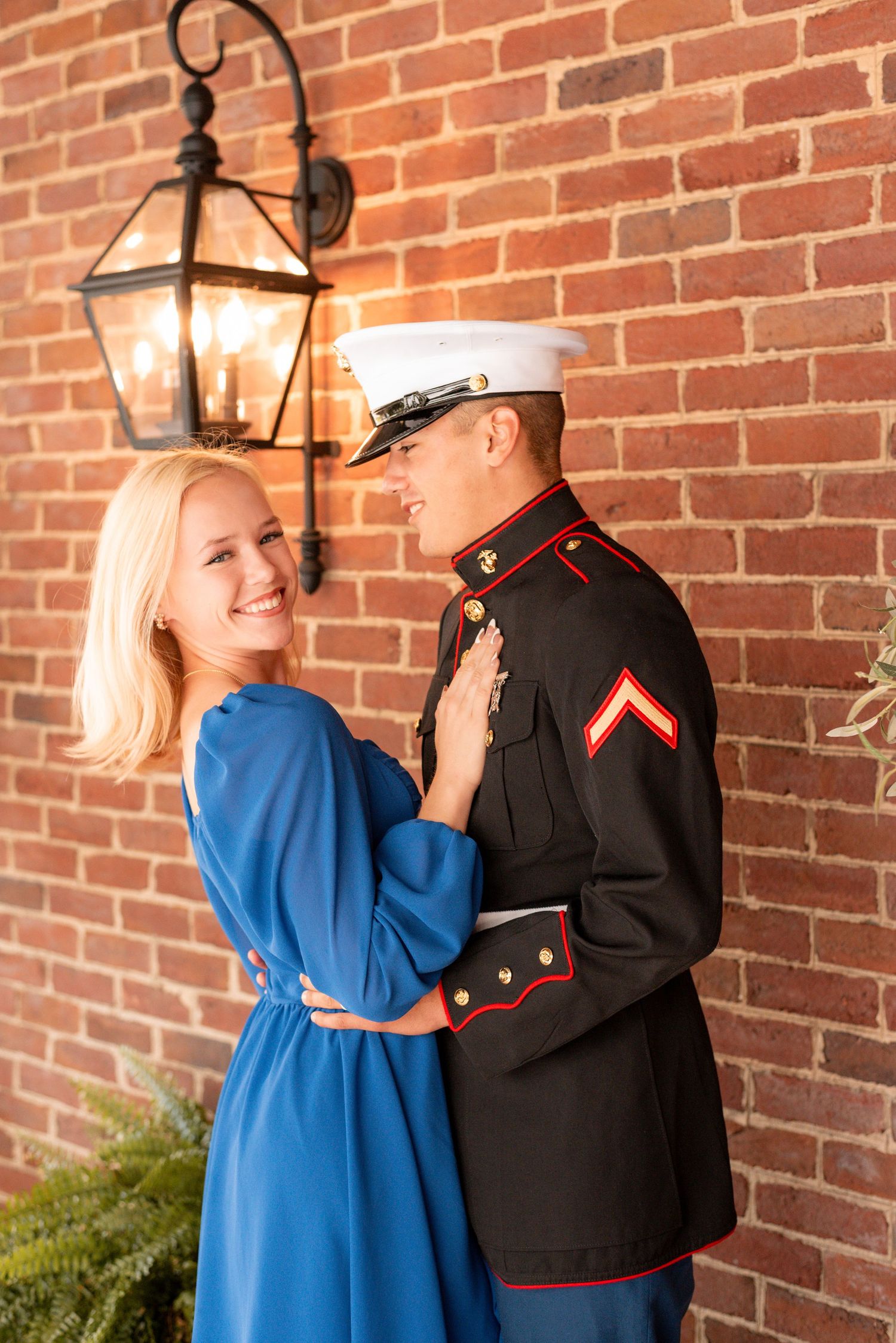 A couple in formal attire embraces against a brick wall with decorative lanterns.
