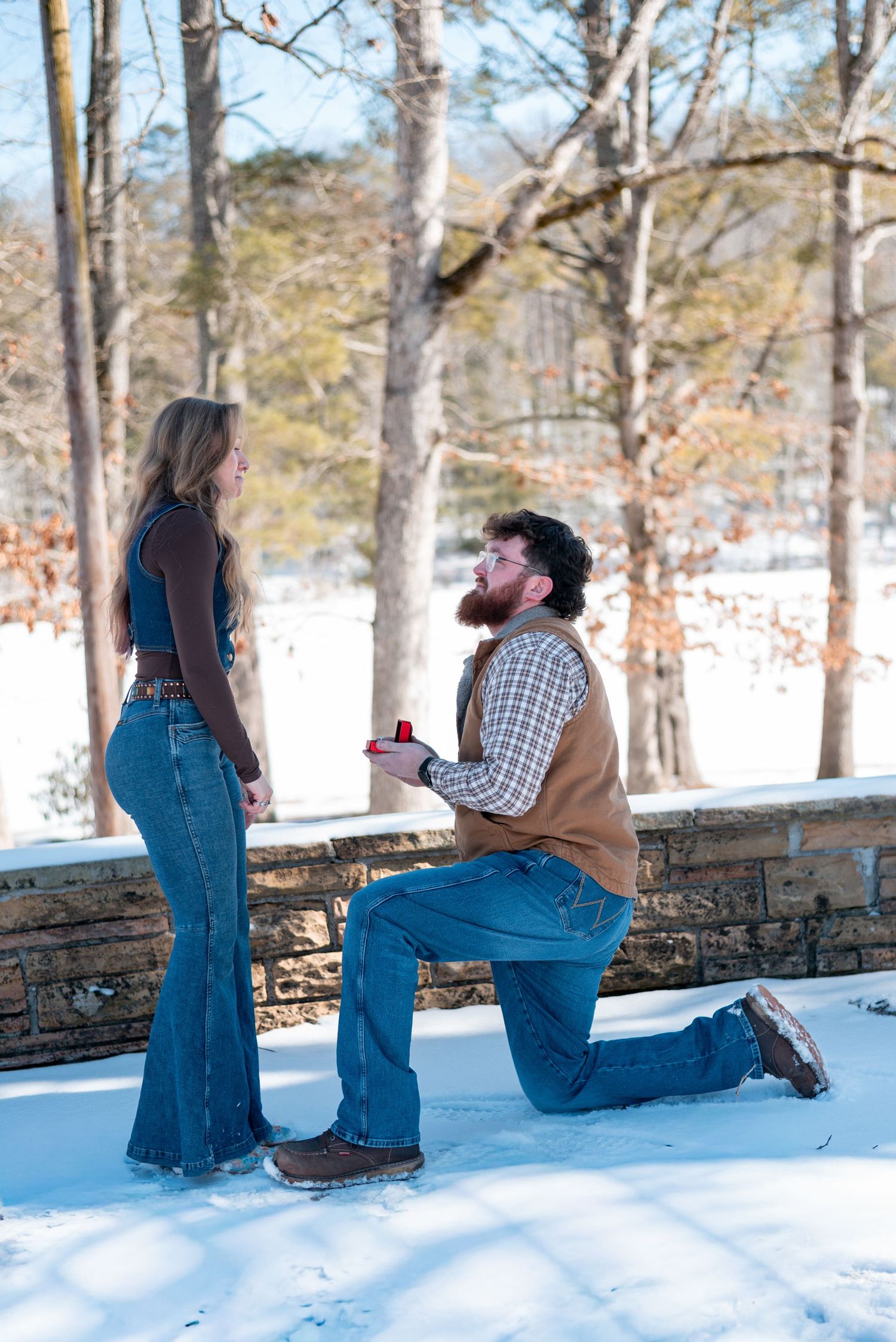 A romantic winter proposal moment on a snow-covered stone wall surrounded by bare trees.