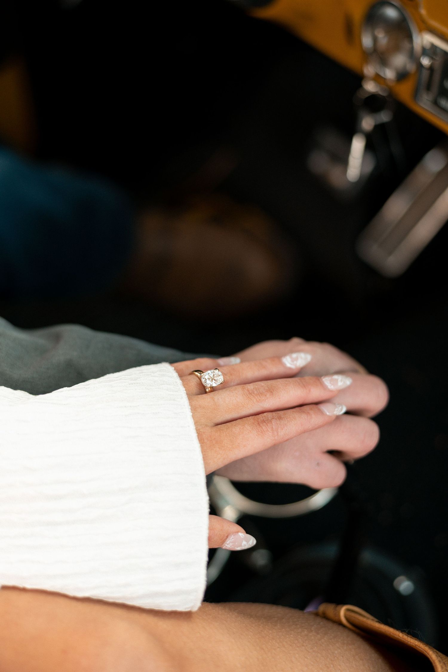 Close-up of hands with white sleeves and engagement ring in dim lighting.