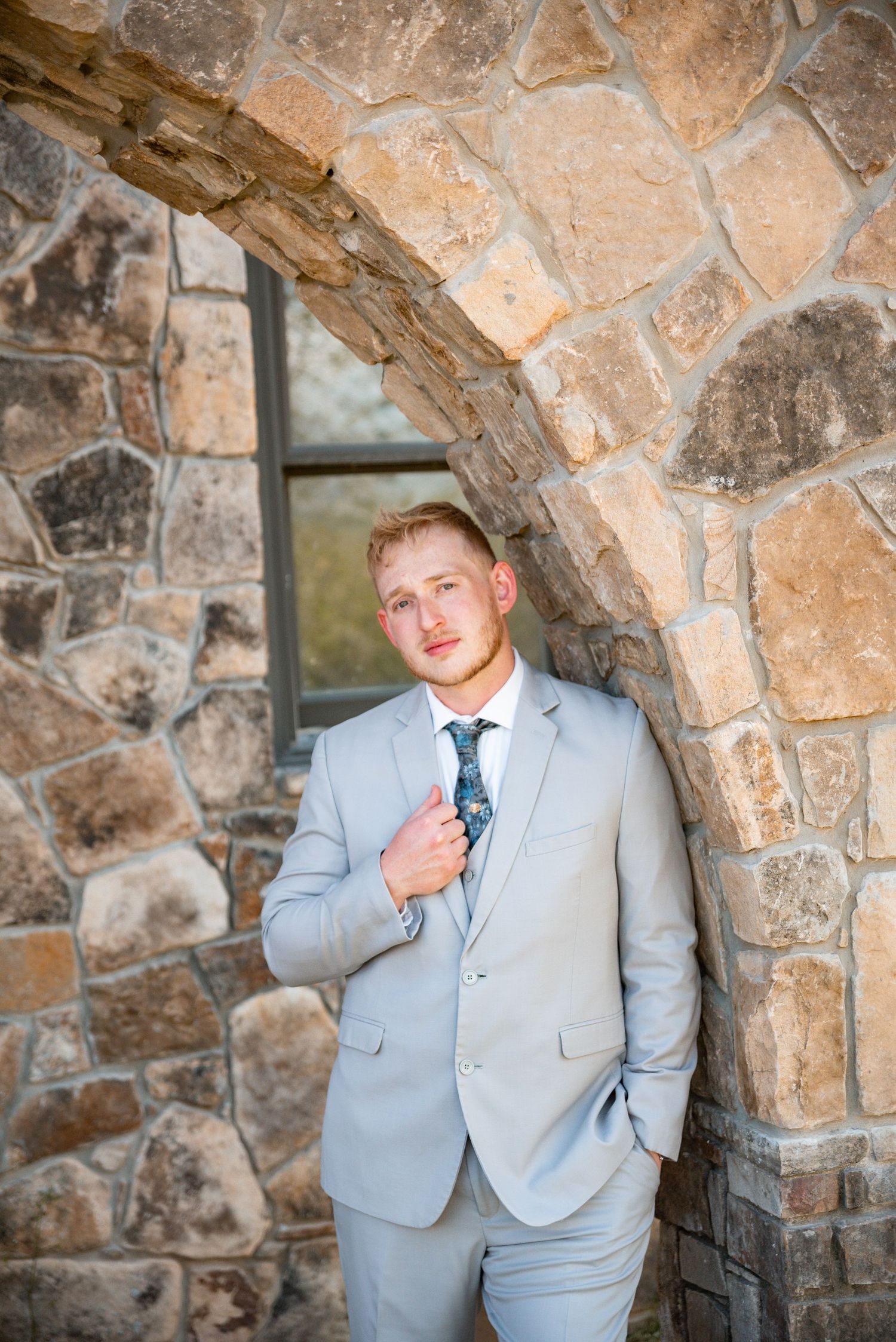 A young man in a light gray suit and floral tie leans against a rustic stone wall under an arch.