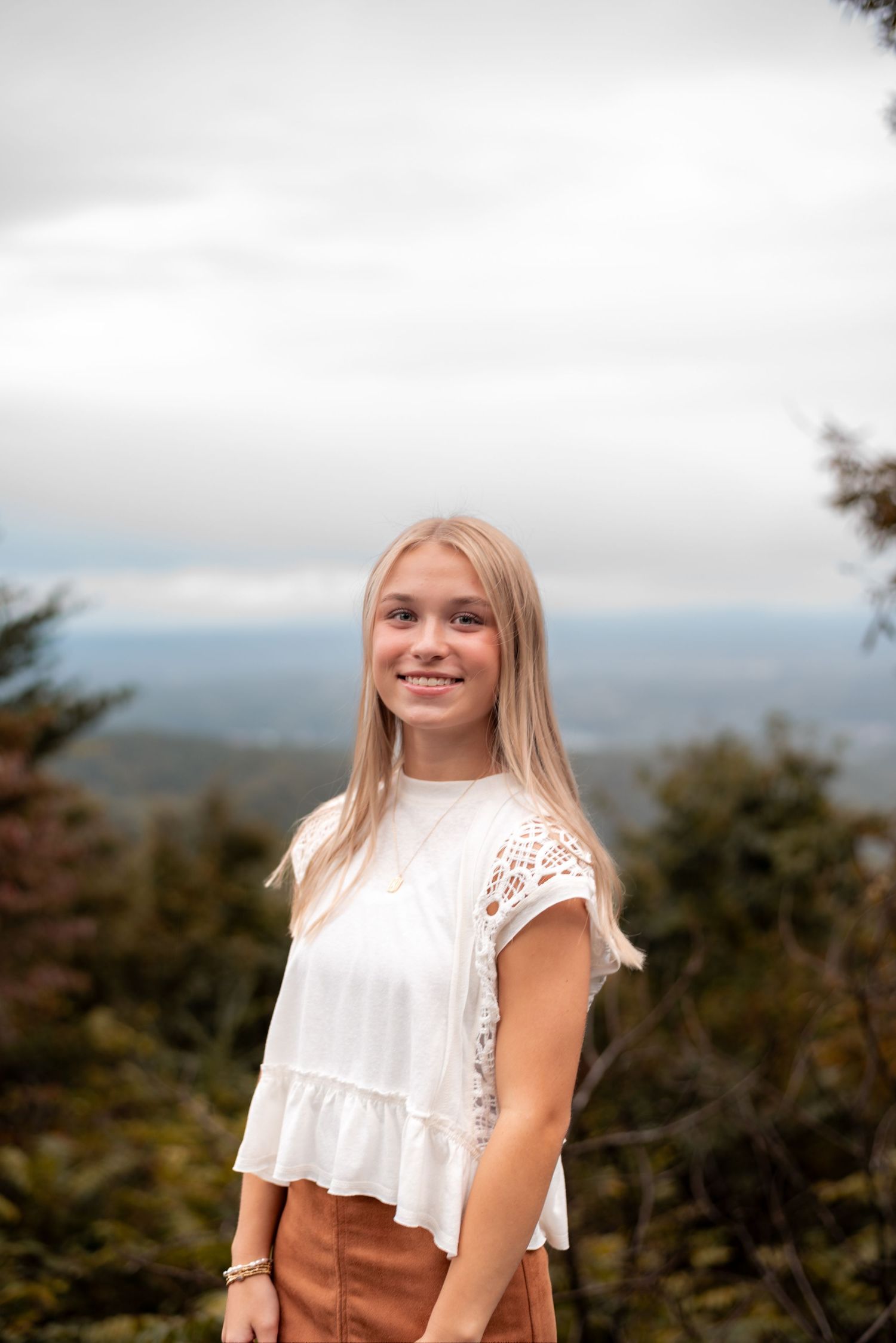 A person in a white lace top poses against a scenic mountain overlook on a cloudy day.