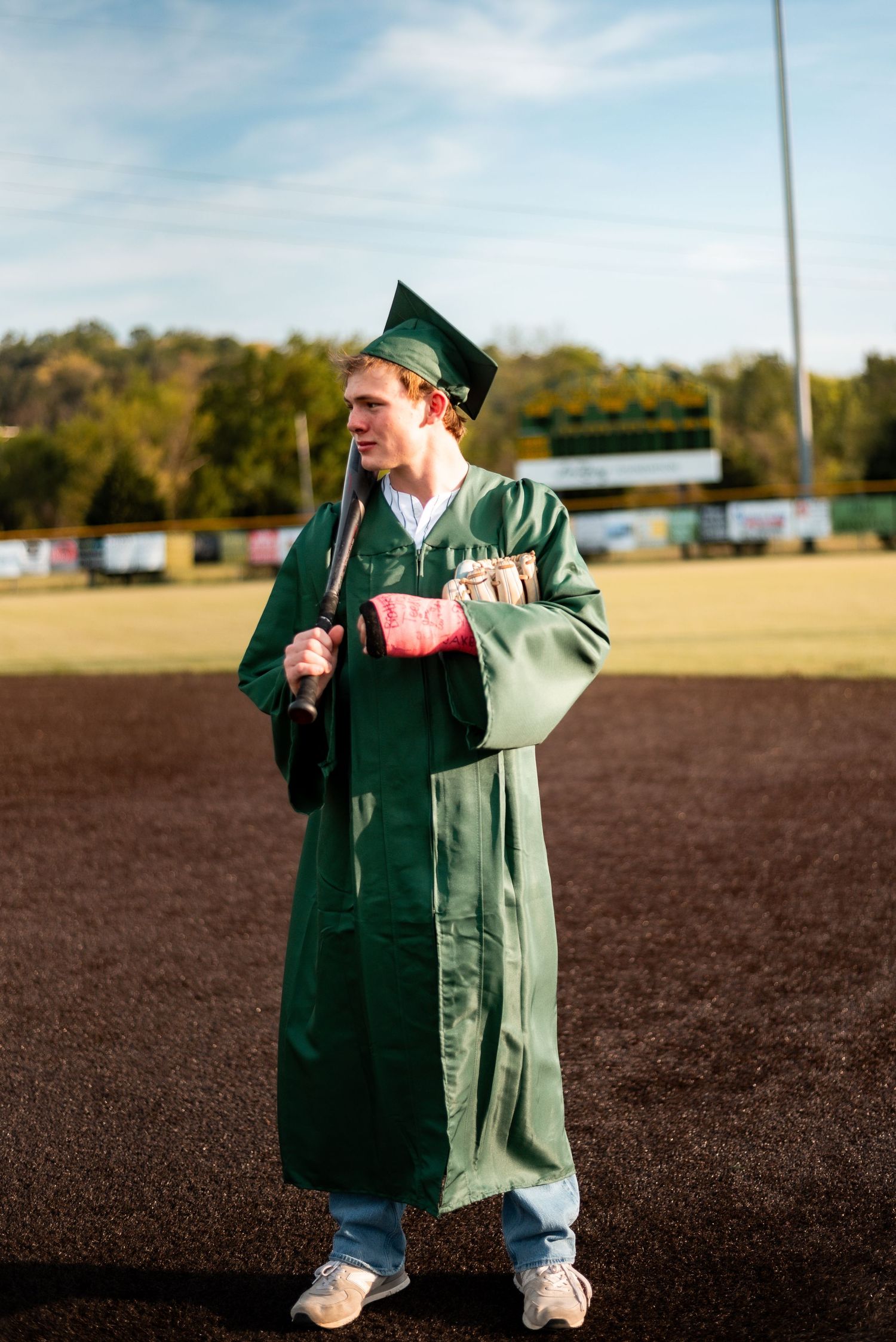 Young man in green graduation cap and gown holding a baseball bat on a field.