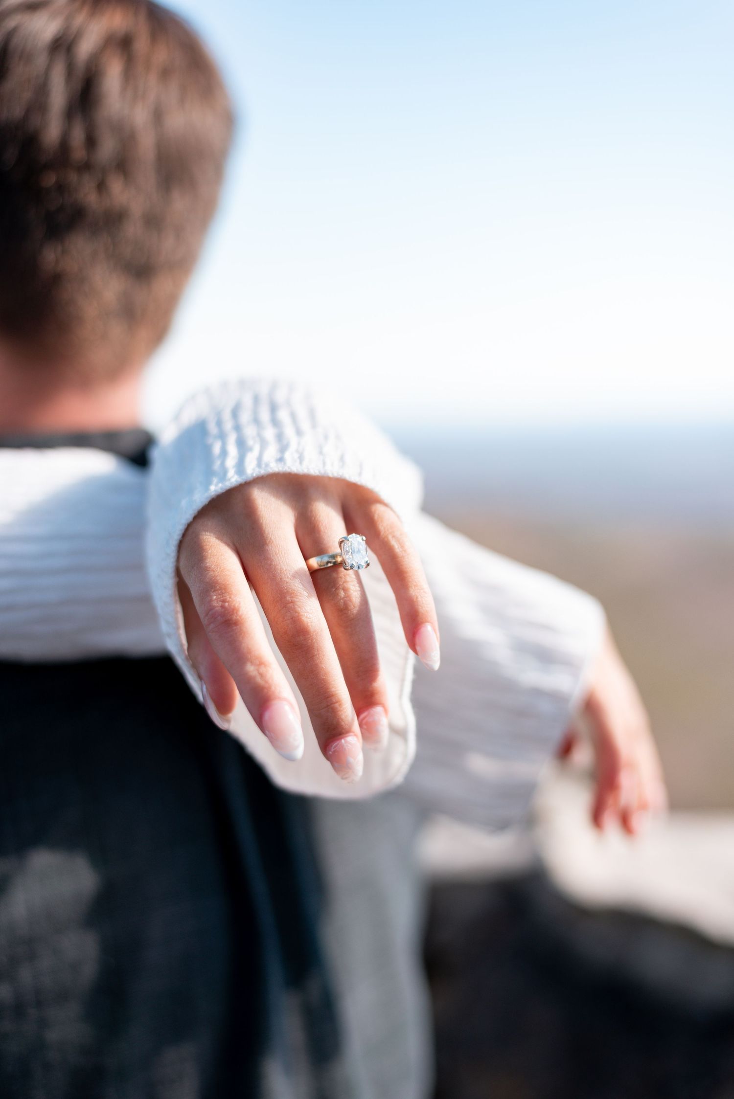 Close-up of hands showing off a sparkling engagement ring against a bright sky background.