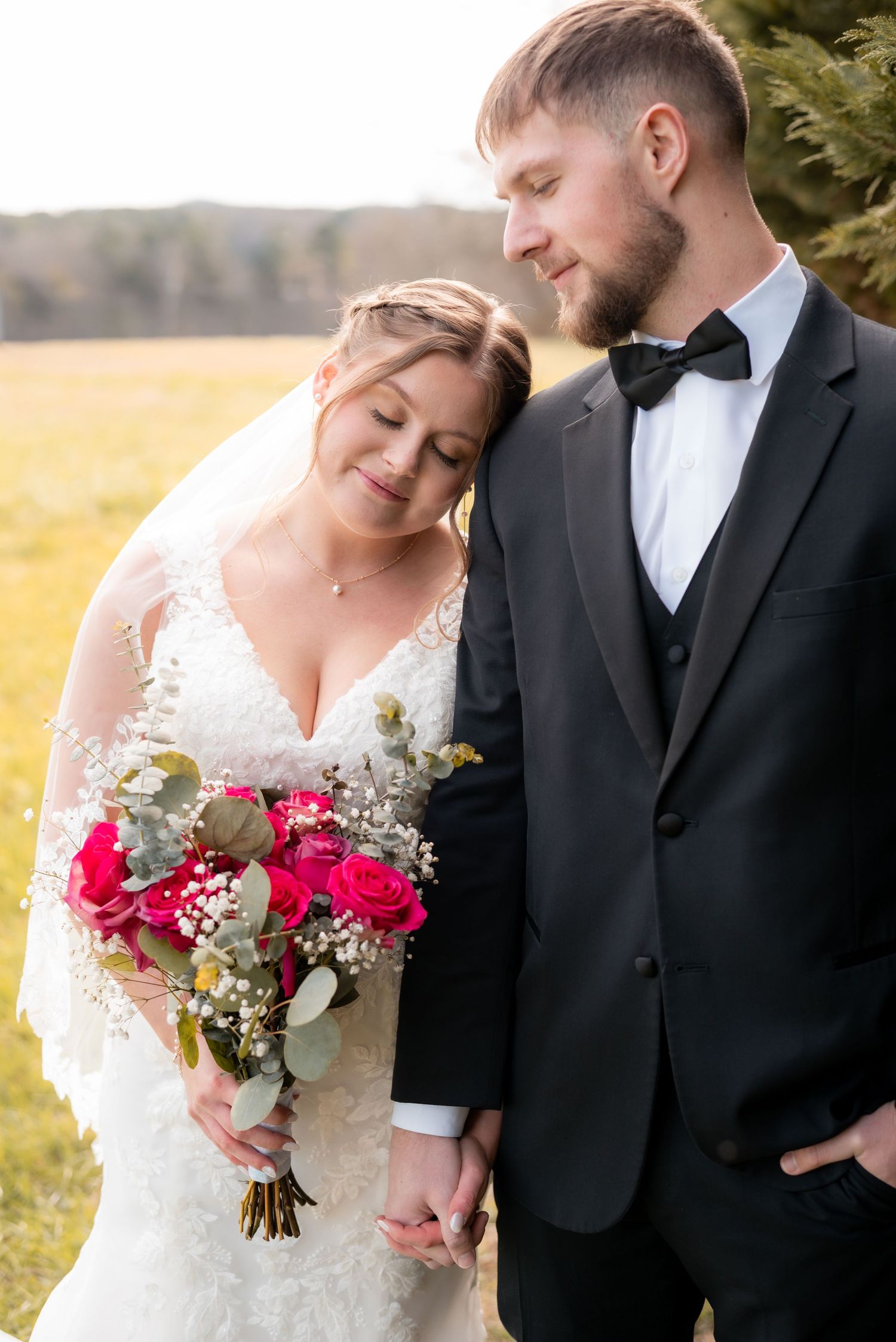 Romantic wedding couple embracing in a sunlit meadow with red and white bouquet.