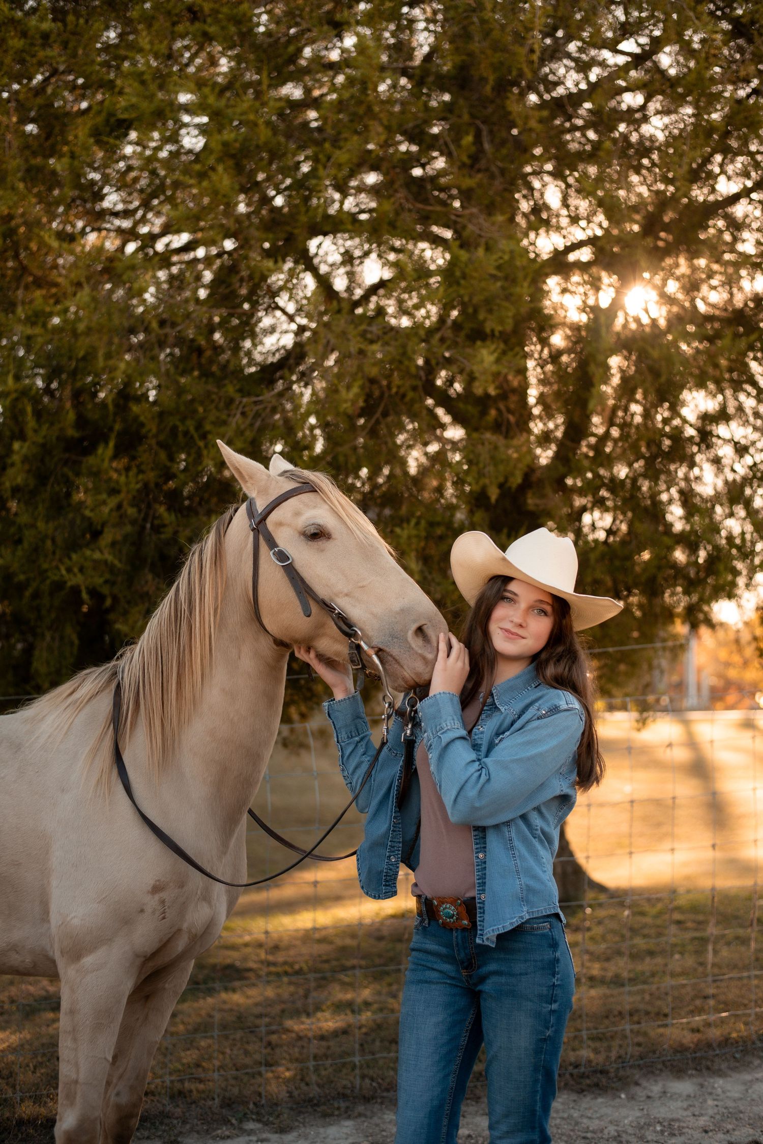 A palomino horse stands with its handler wearing denim and a cowboy hat at sunset.