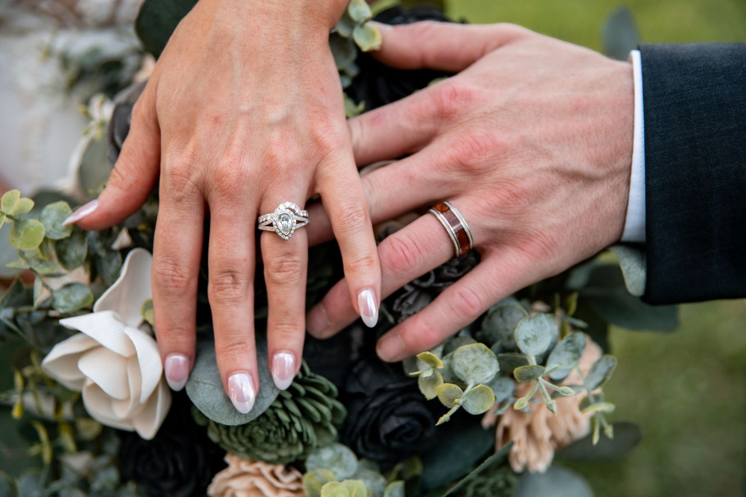 Bride and groom displaying wedding rings on a floral bouquet with white and dark blooms.