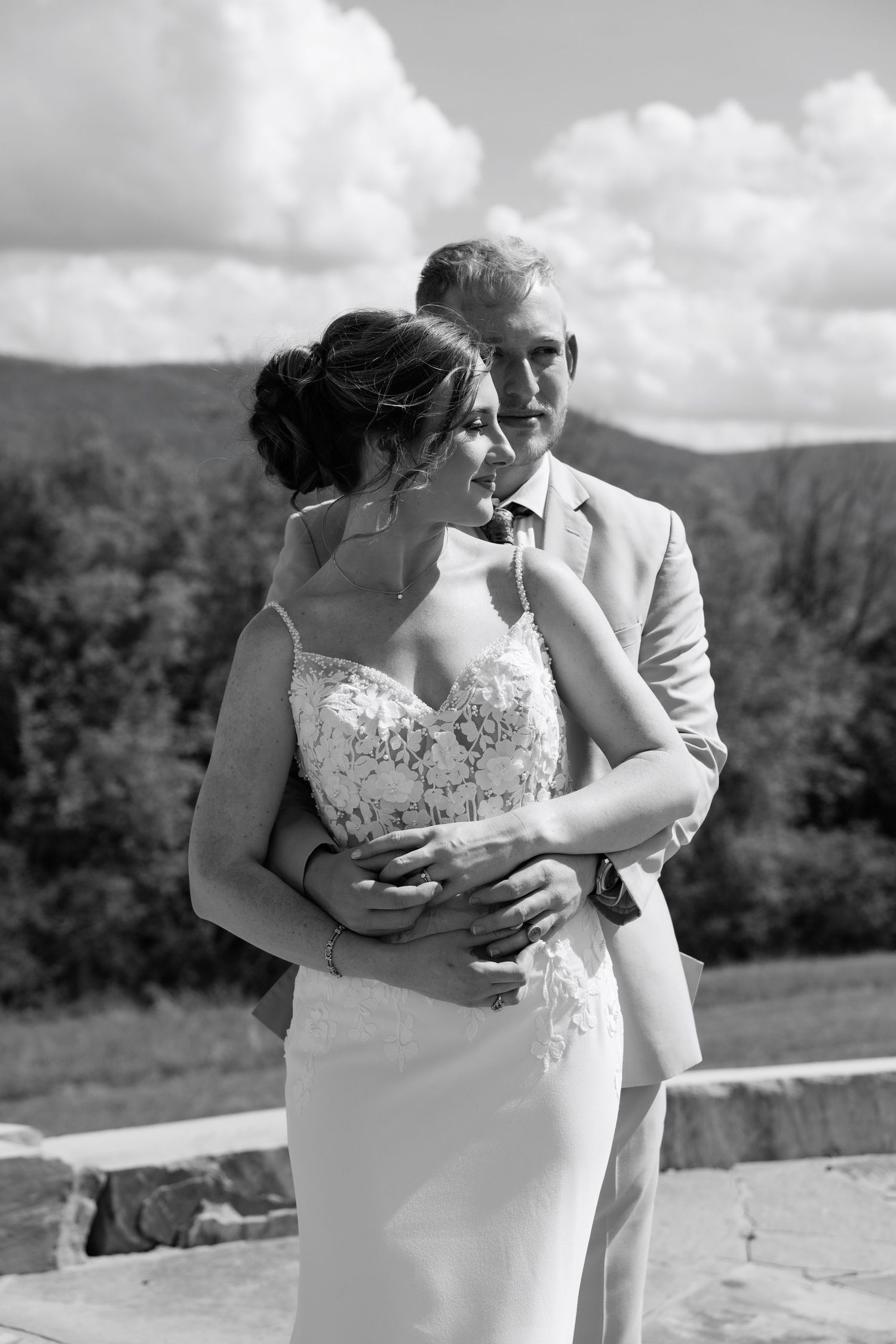 Black and white photo of a bride and groom embracing outdoors with mountain scenery and clouds behind them.