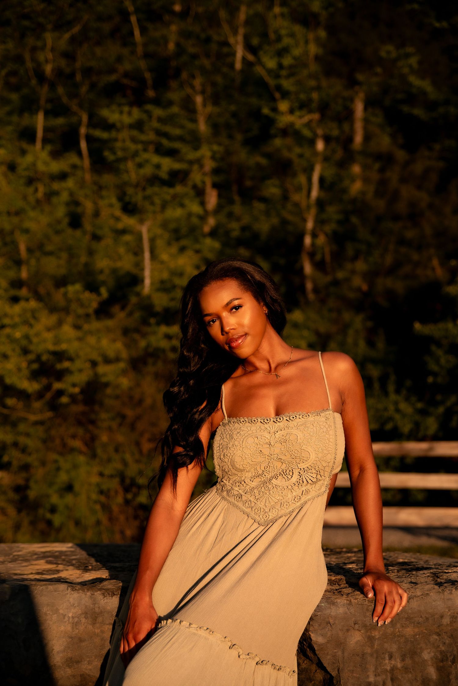 A woman in a white lace sundress poses gracefully against a stone railing with lush green trees in golden sunset light.