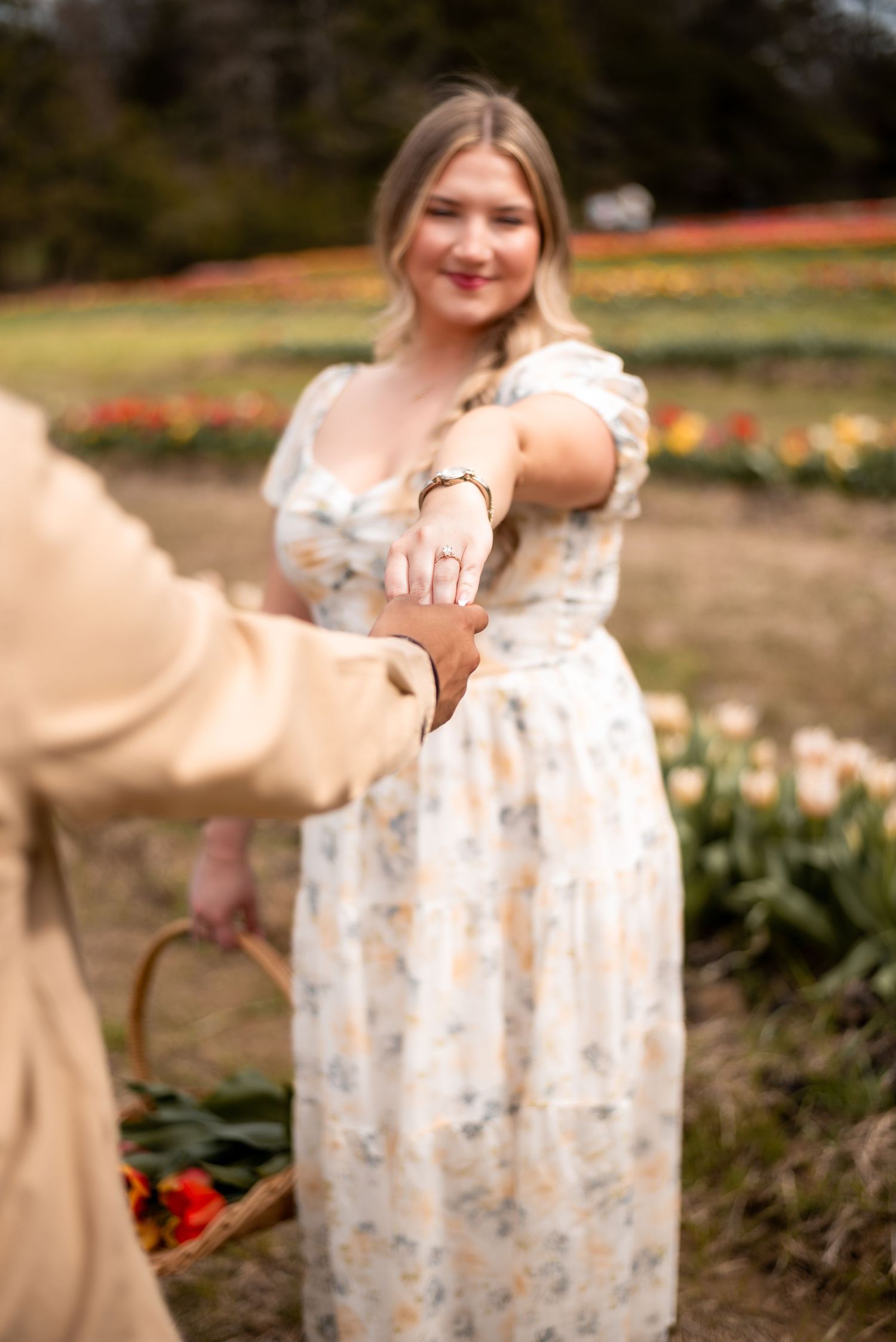 A smiling blonde woman in a floral dress holds hands with a partner in a tulip field, showcasing an engagement ring.