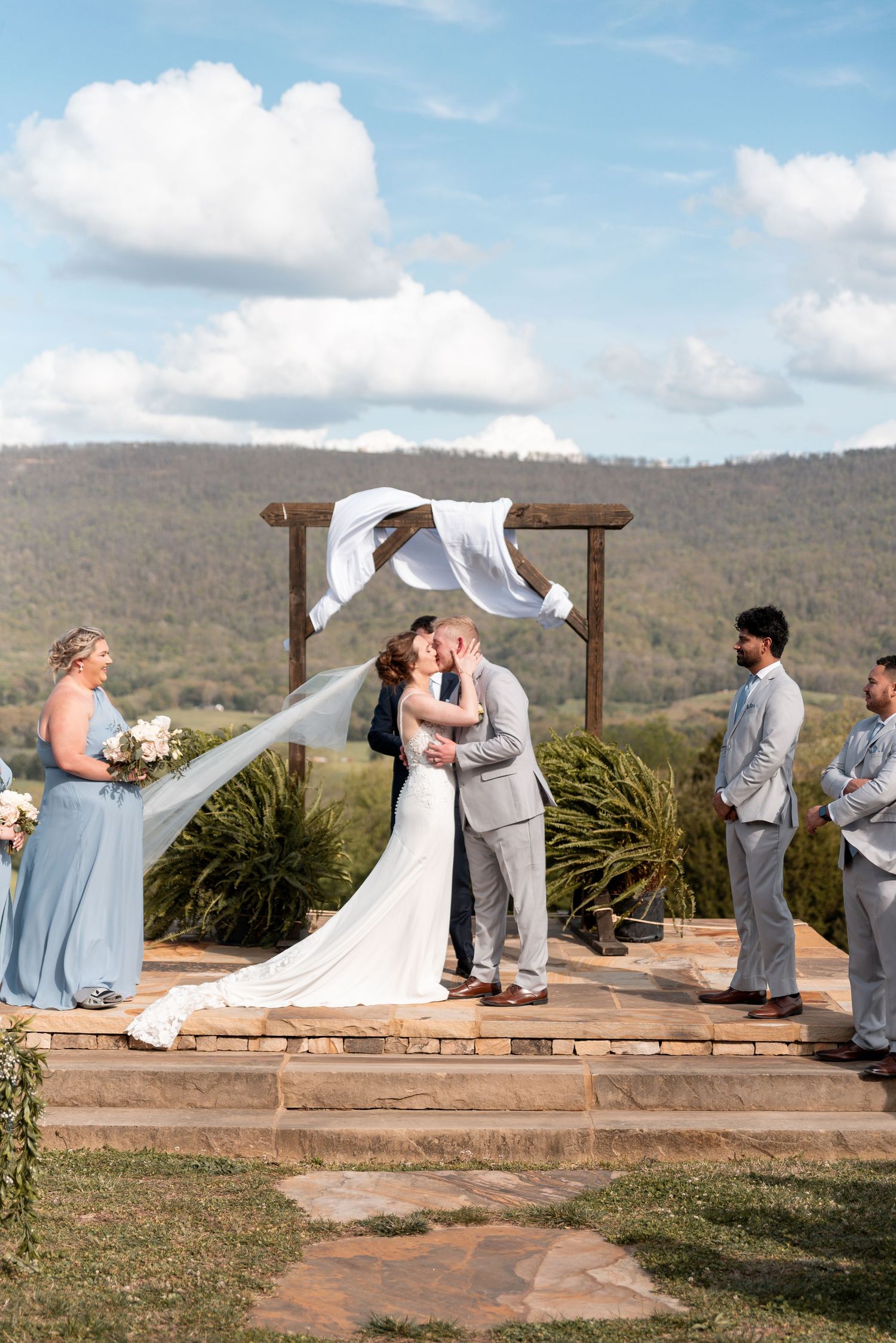 Bride and groom share first kiss at outdoor wedding ceremony with mountain views and windswept veil.