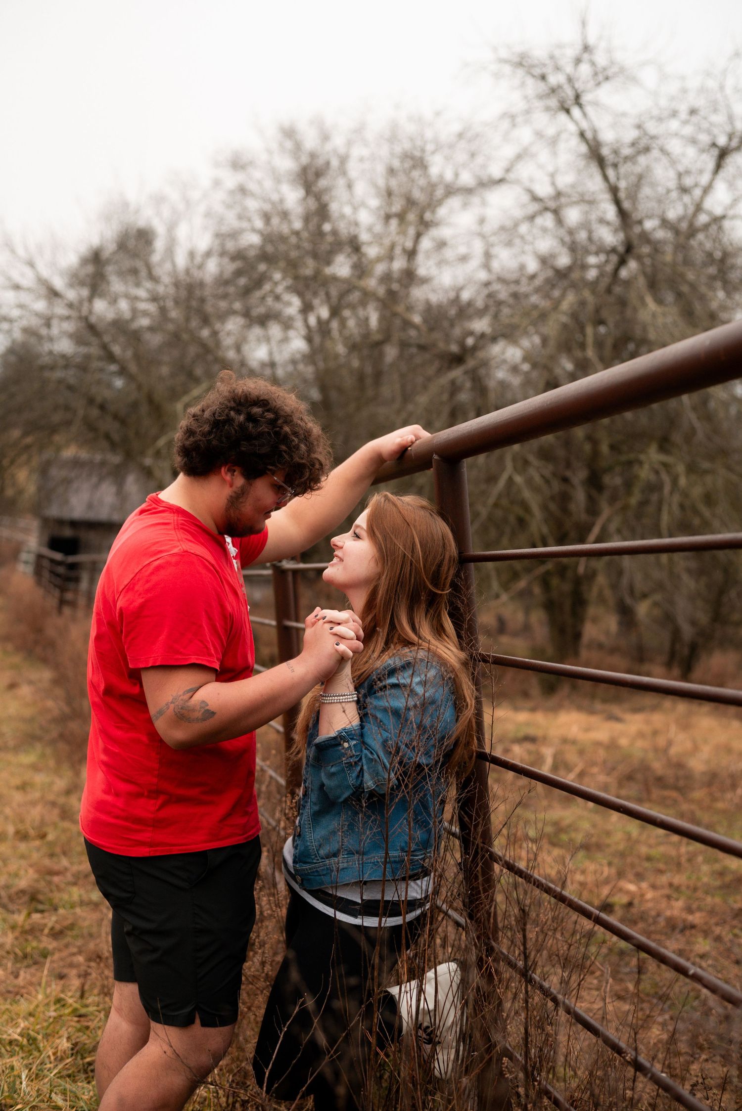 Two people share a playful moment leaning against a rustic fence in a rural setting.