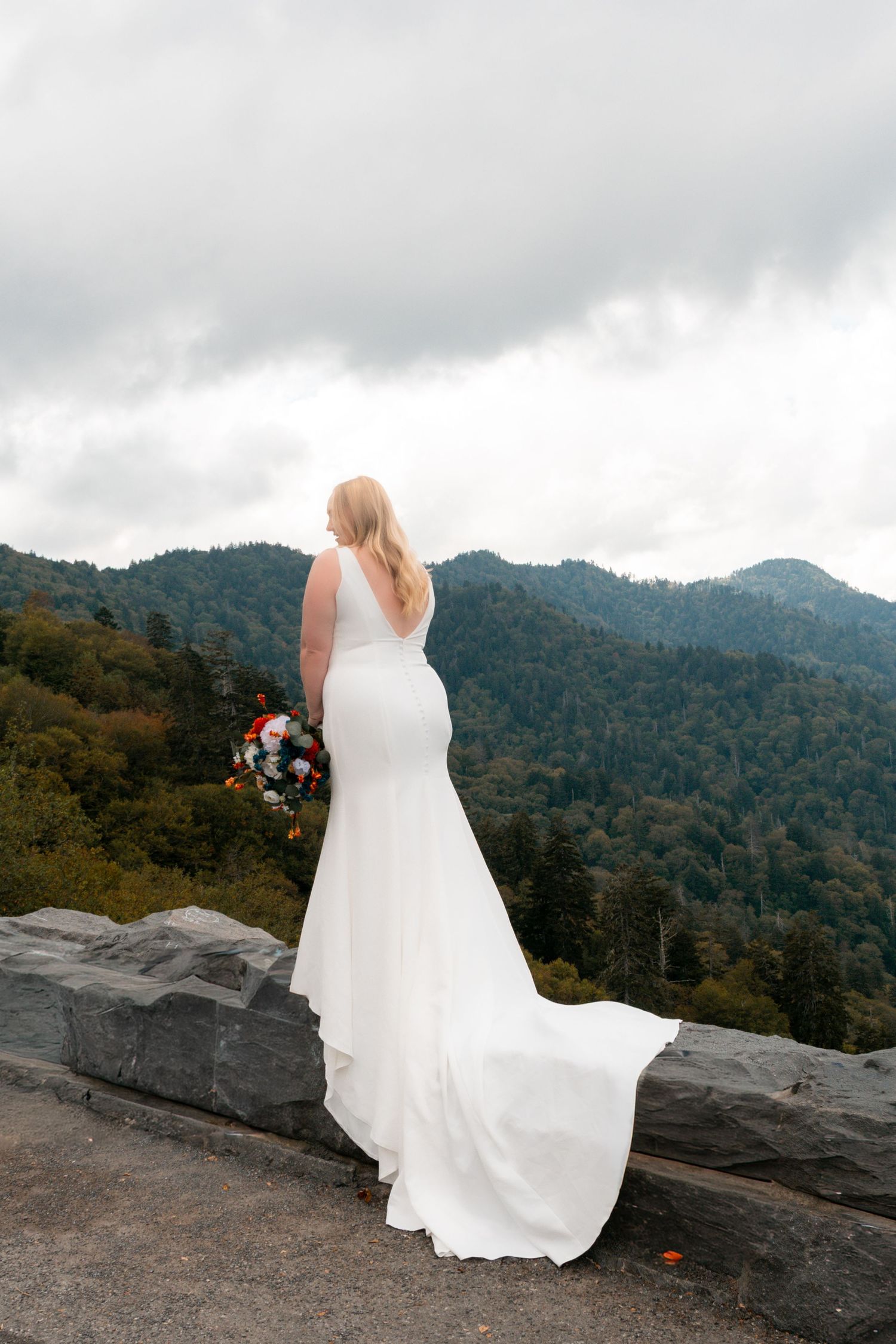 Bride in flowing white dress stands on mountain overlook with dramatic cloudy backdrop.