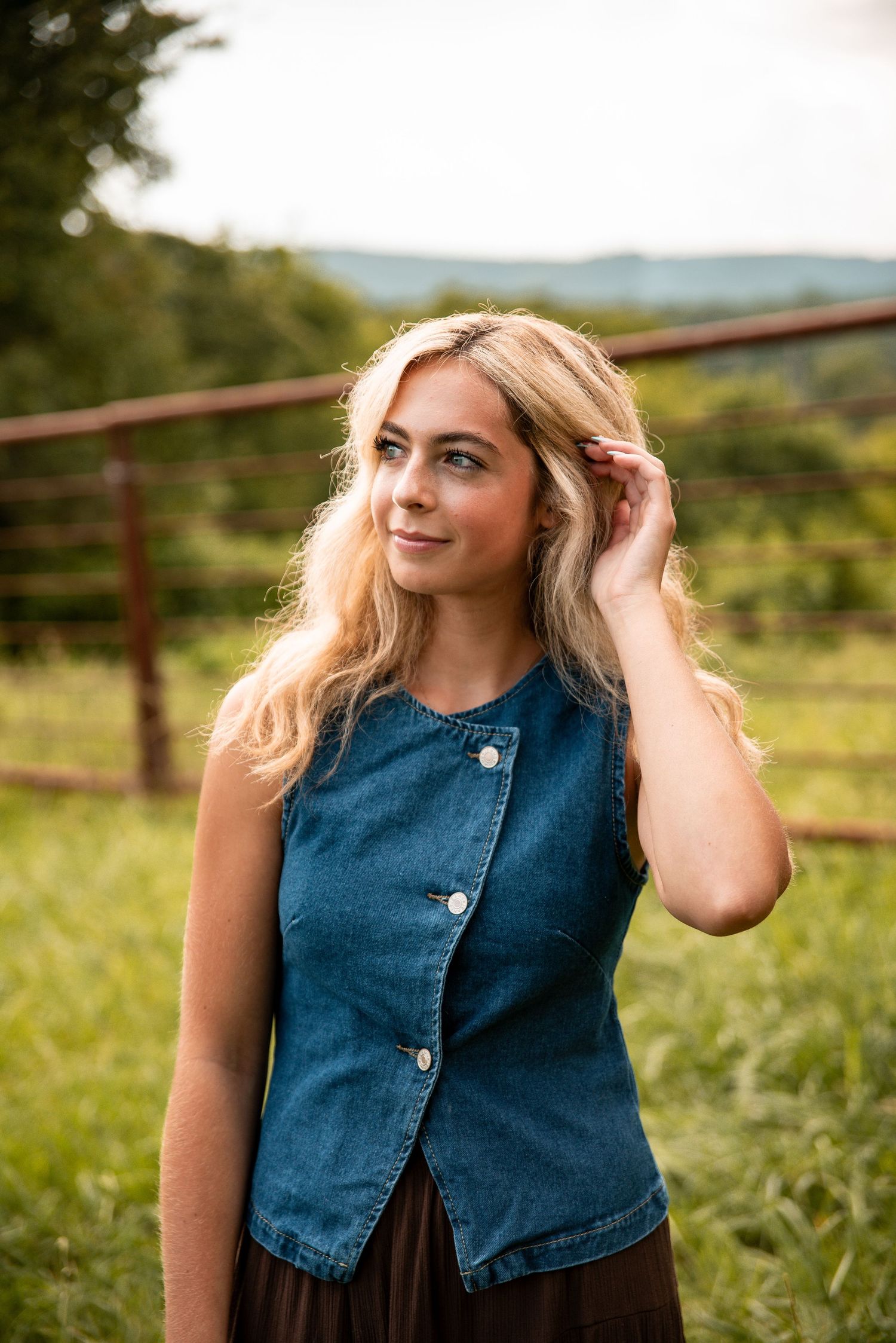 Blonde woman in denim vest standing outdoors in green field with rustic fence and rolling hills.