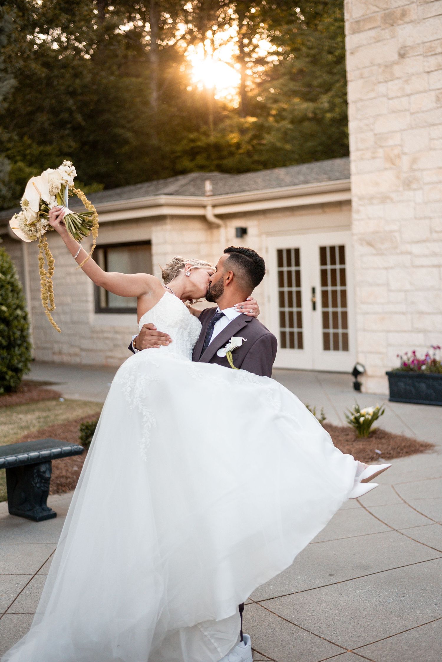 Groom lifts bride in white gown as they kiss outdoors at sunset, bride holding bouquet aloft near stone building.