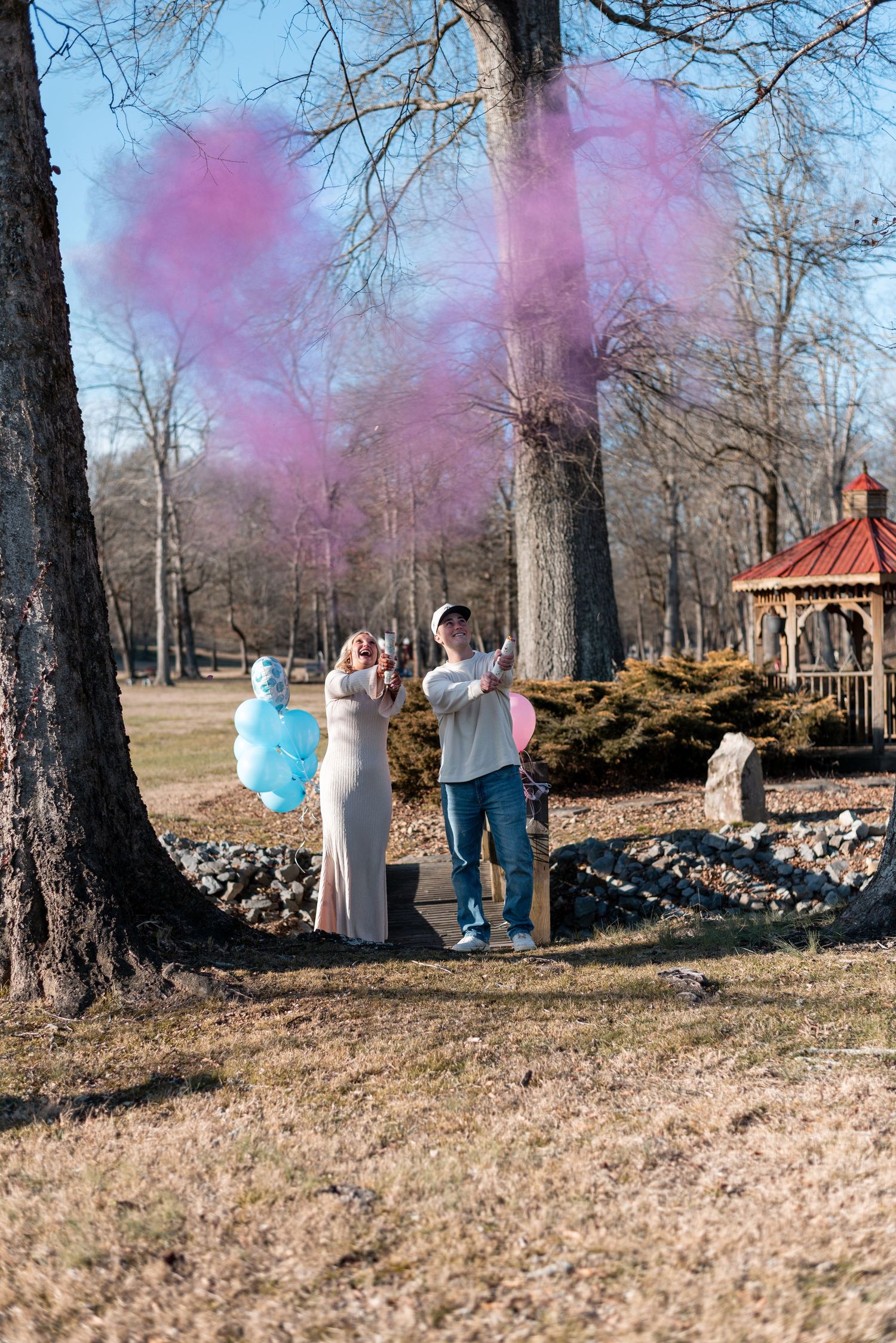 Group celebration with pink smoke bombs in outdoor park setting among bare trees.