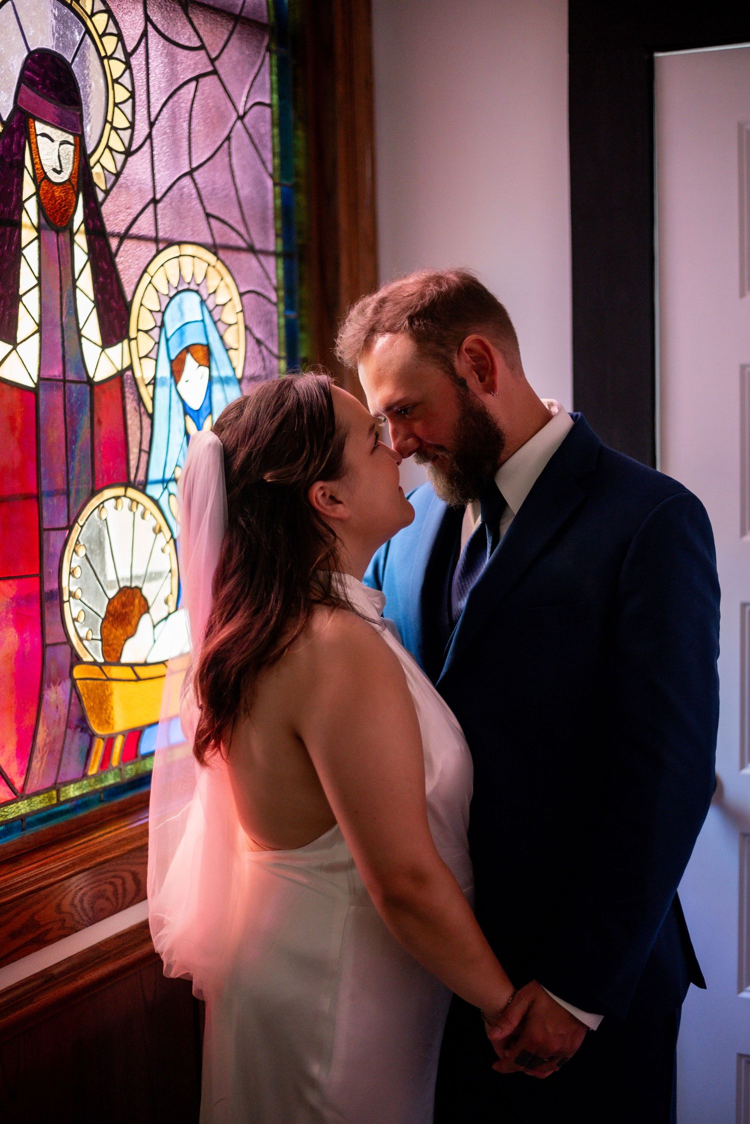 Couple shares romantic moment in front of colorful stained glass window, bride in white gown and groom in navy suit.