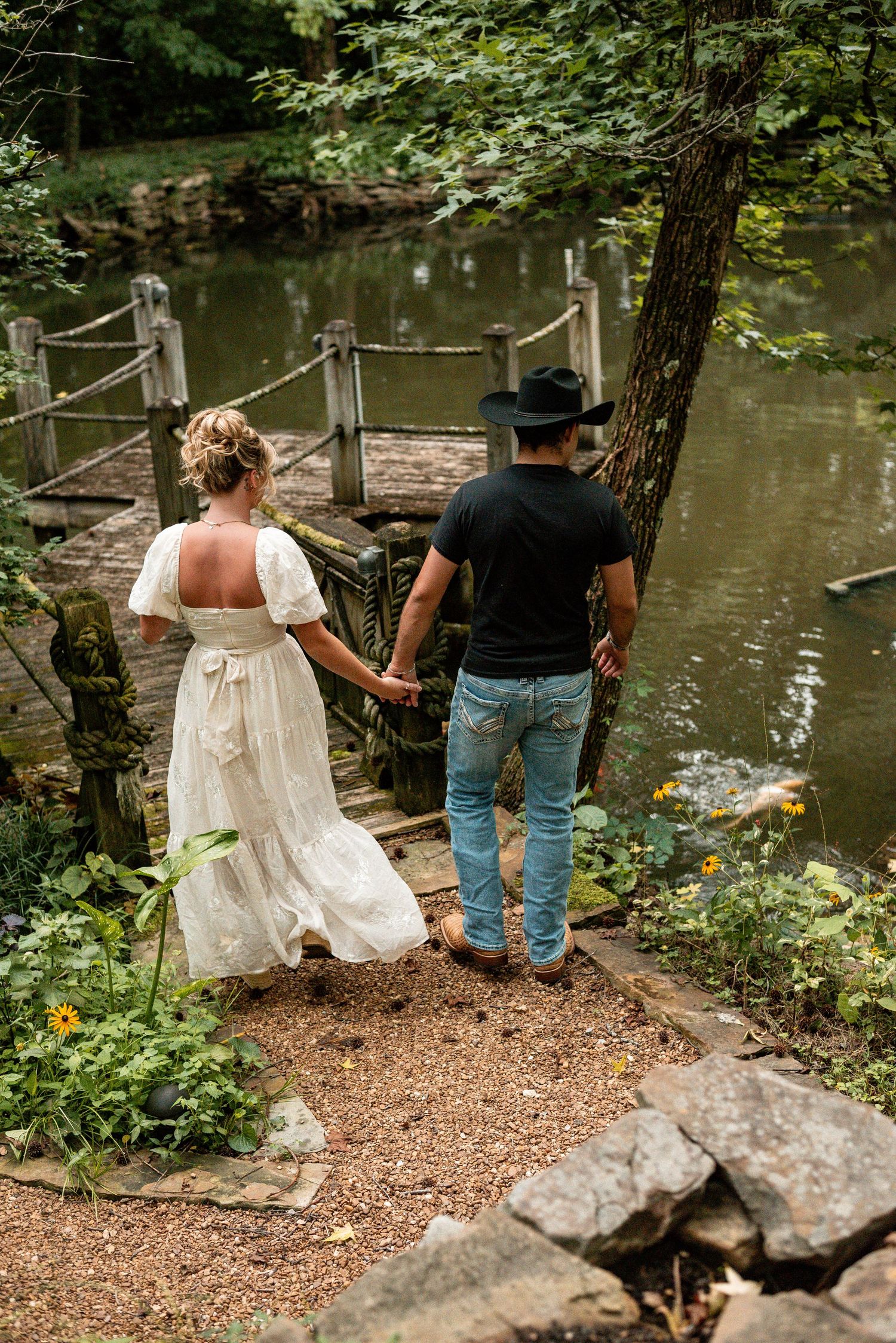 A couple in wedding attire holds hands walking toward a rustic dock by a serene pond surrounded by lush greenery.