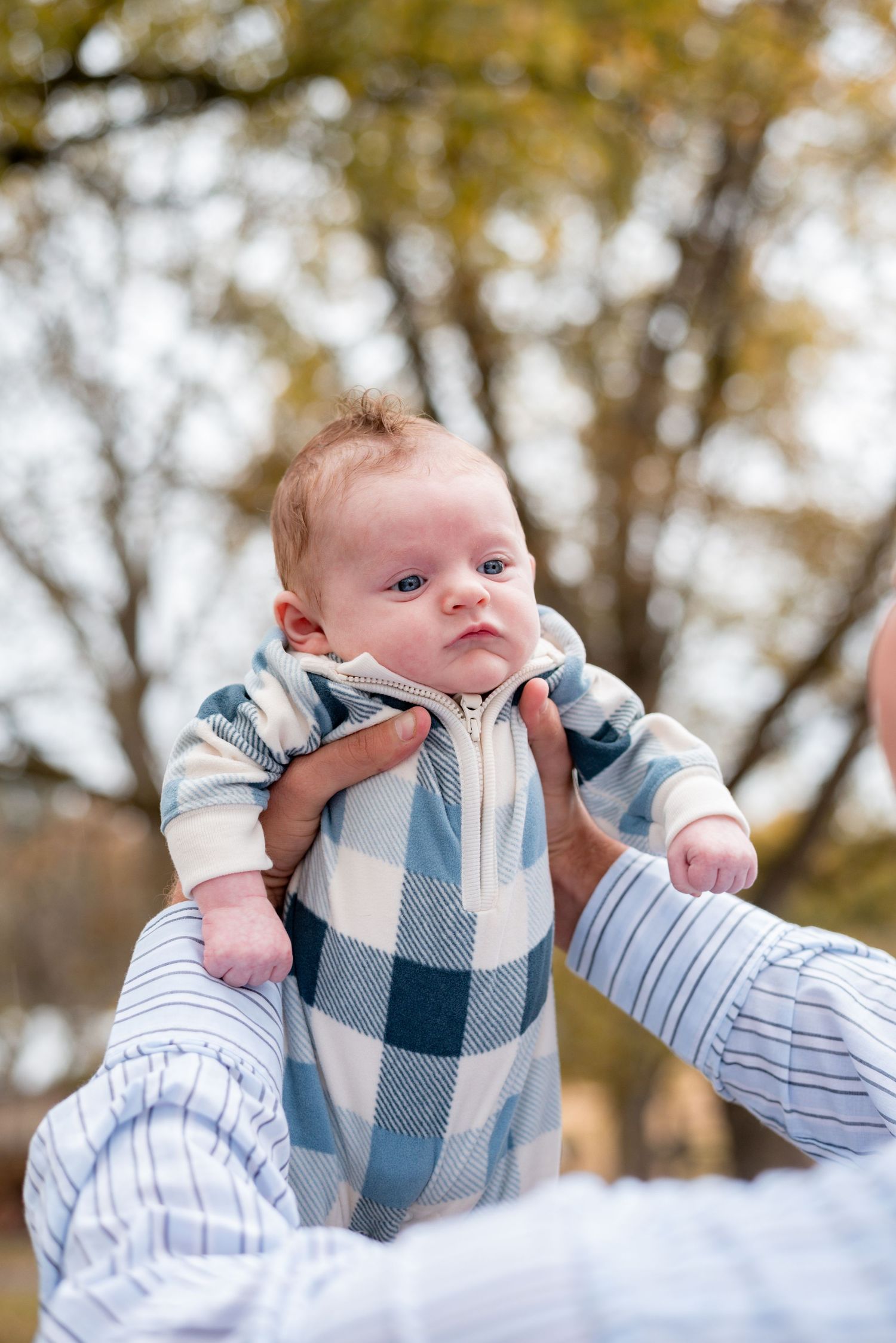 Baby wearing blue plaid vest and white shirt looking up with curious expression outdoors.