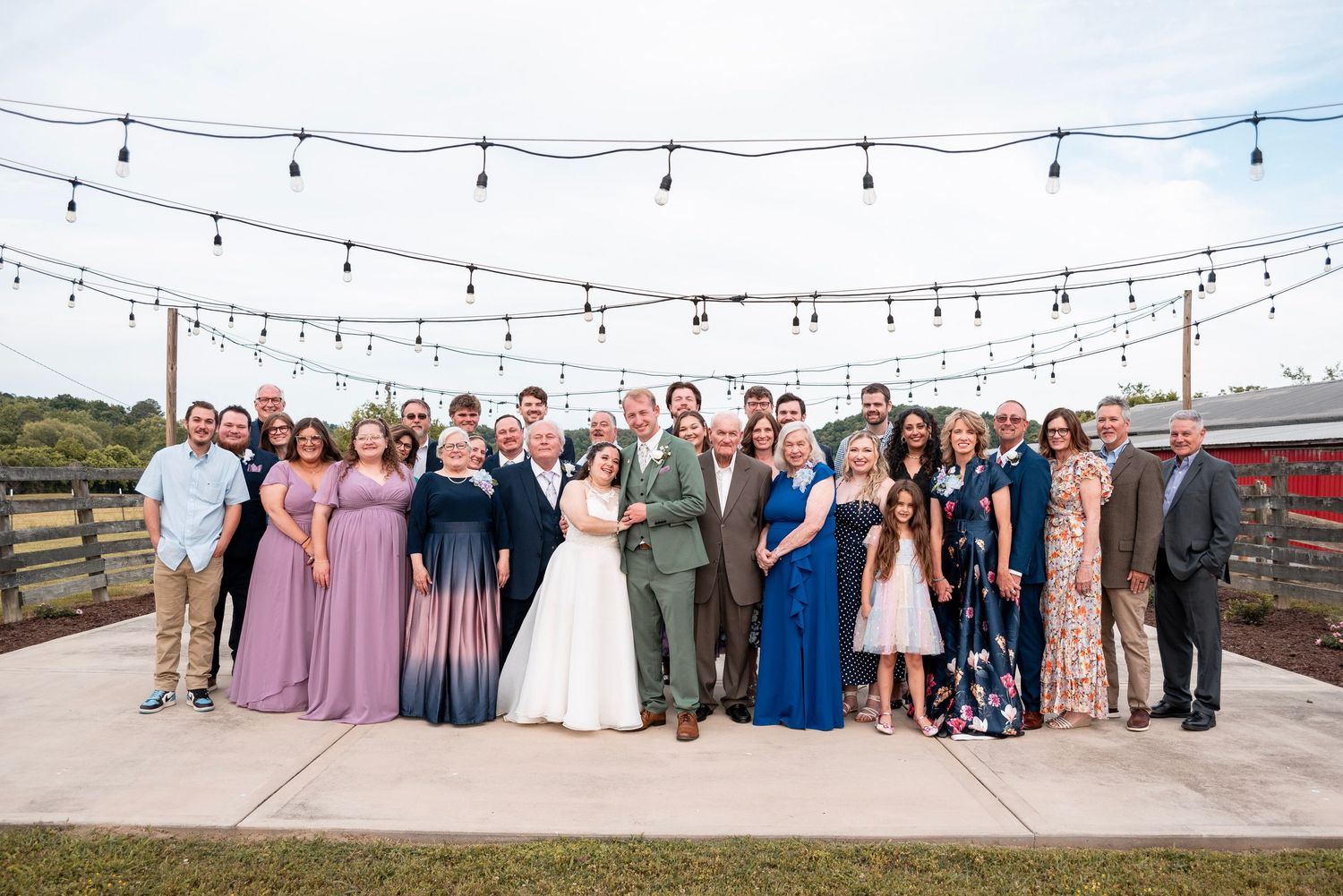Large wedding party group photo outdoors under string lights, featuring bride and groom surrounded by family and friends.
