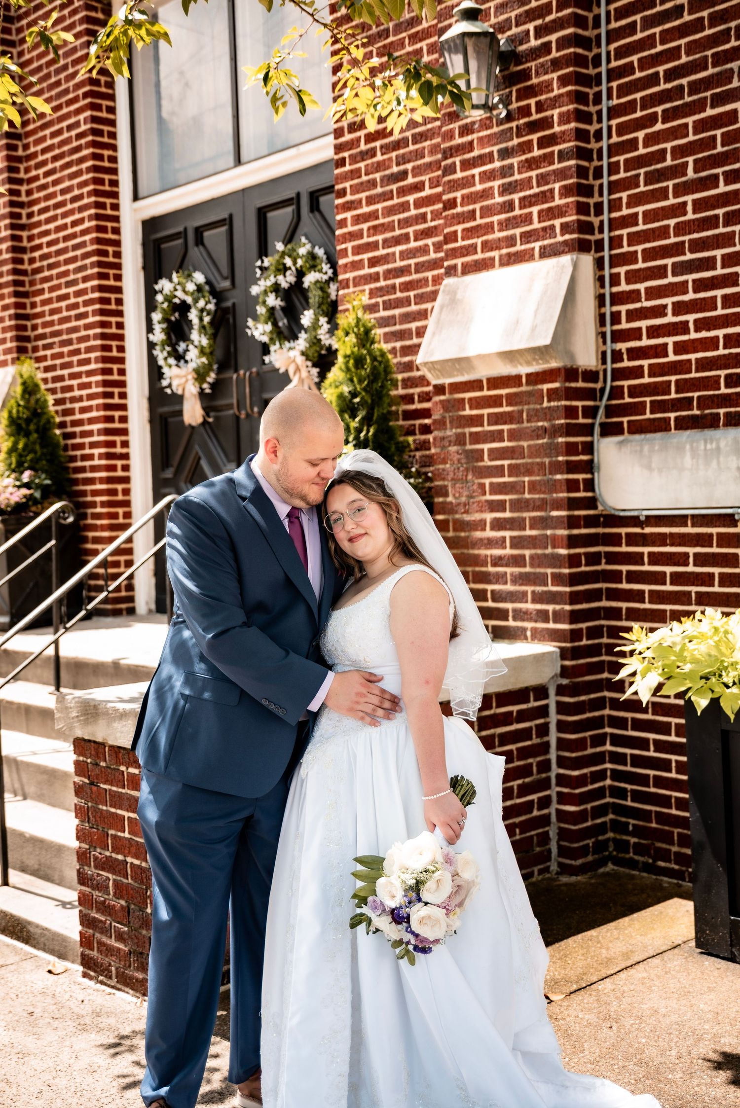 Newlyweds embrace tenderly outside a brick church, bride in white gown holding bouquet, groom in navy suit.
