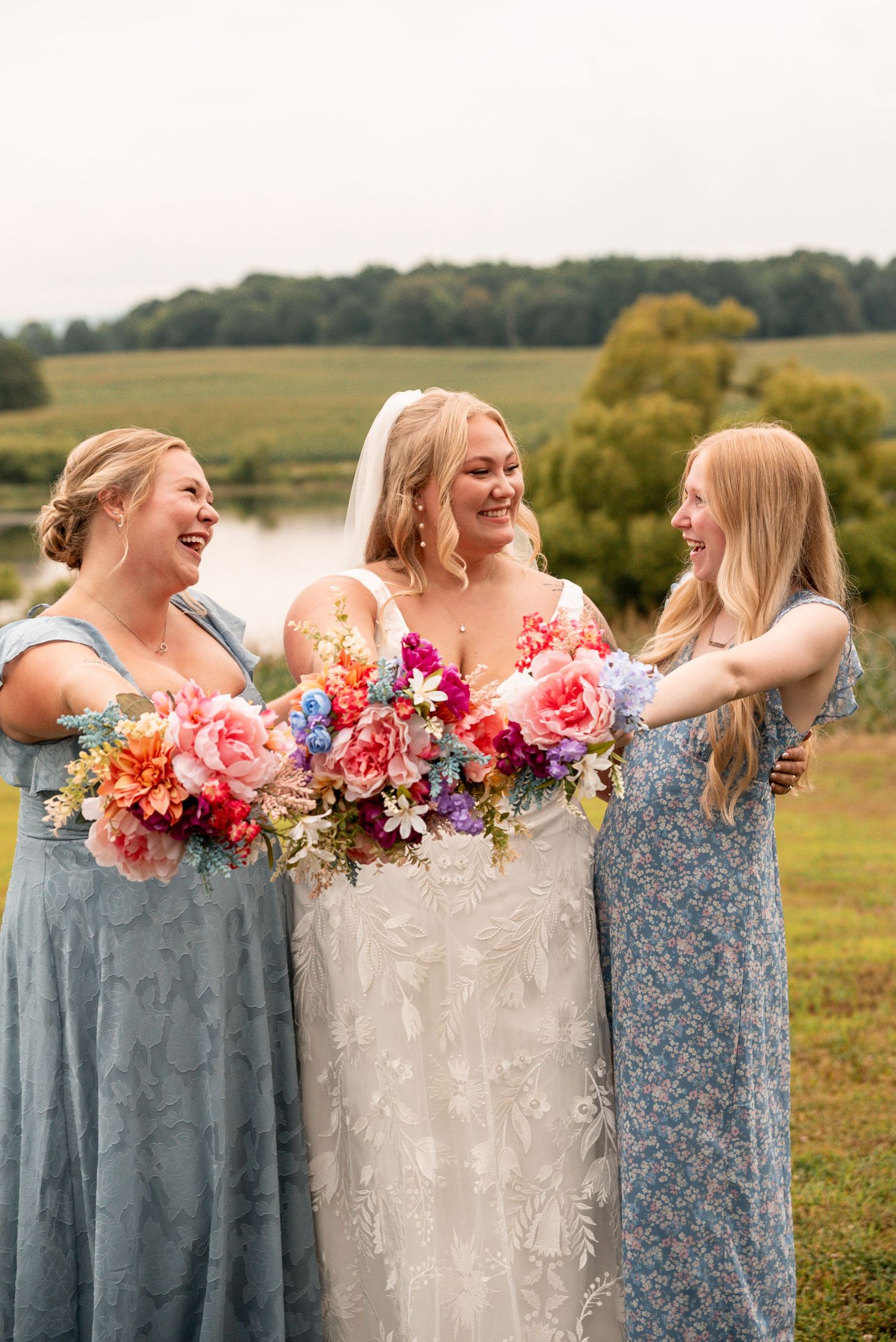 Bride in lace gown laughing with two bridesmaids in blue dresses holding colorful floral bouquets outdoors.