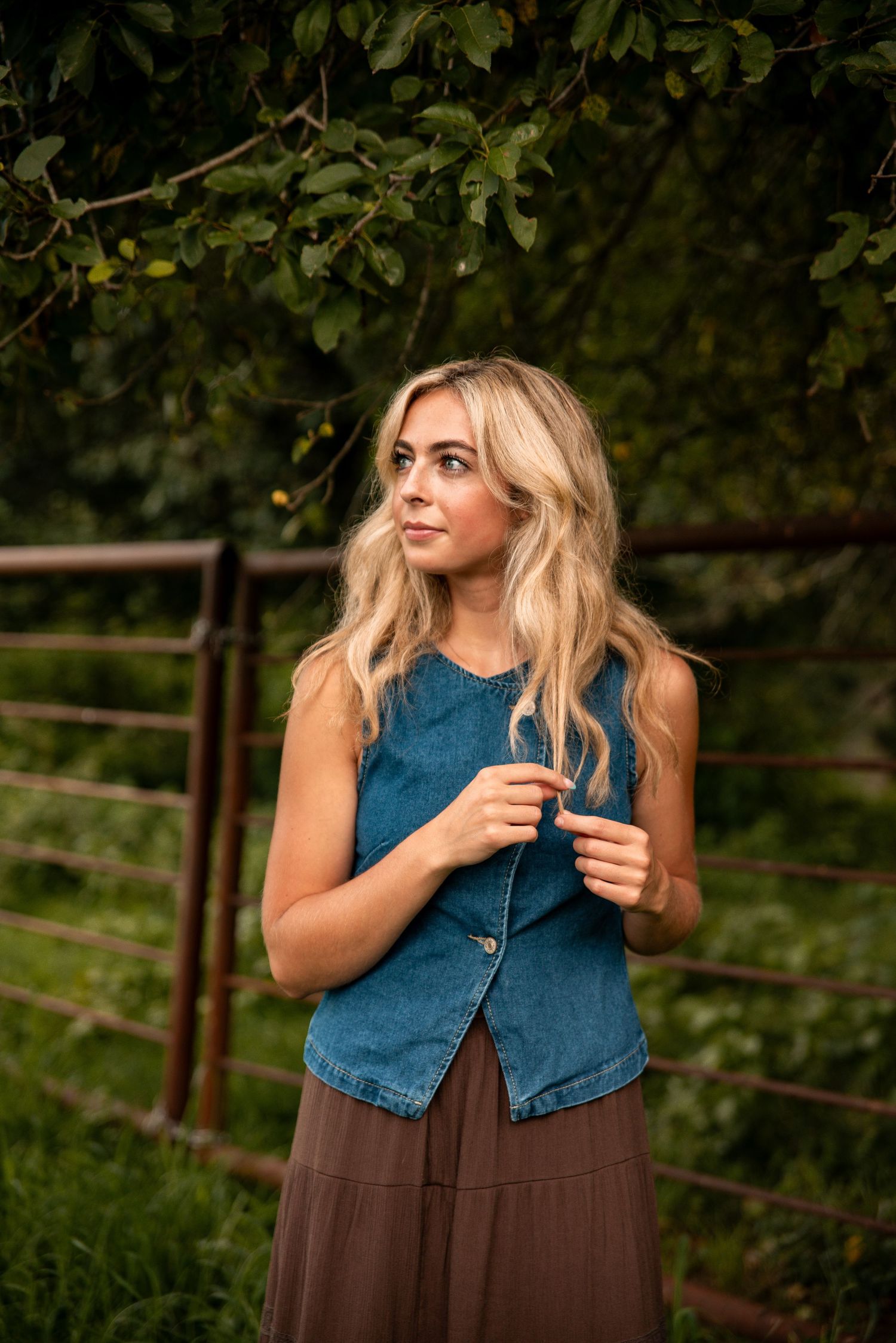 Young blonde woman in denim vest and brown skirt standing outdoors near a rustic metal gate surrounded by lush greenery.