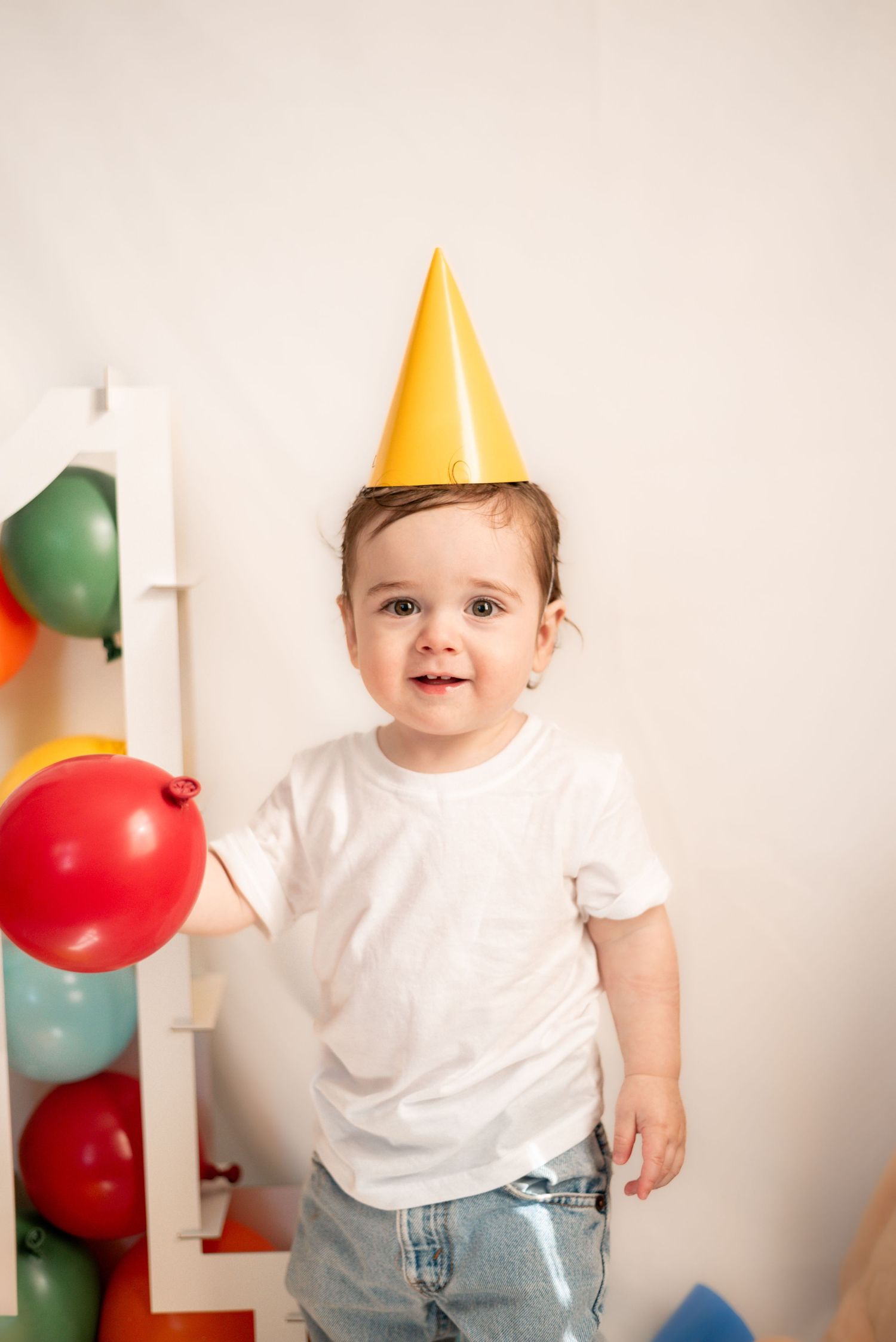 A cheerful toddler in a yellow party hat and white t-shirt poses with colorful balloons.