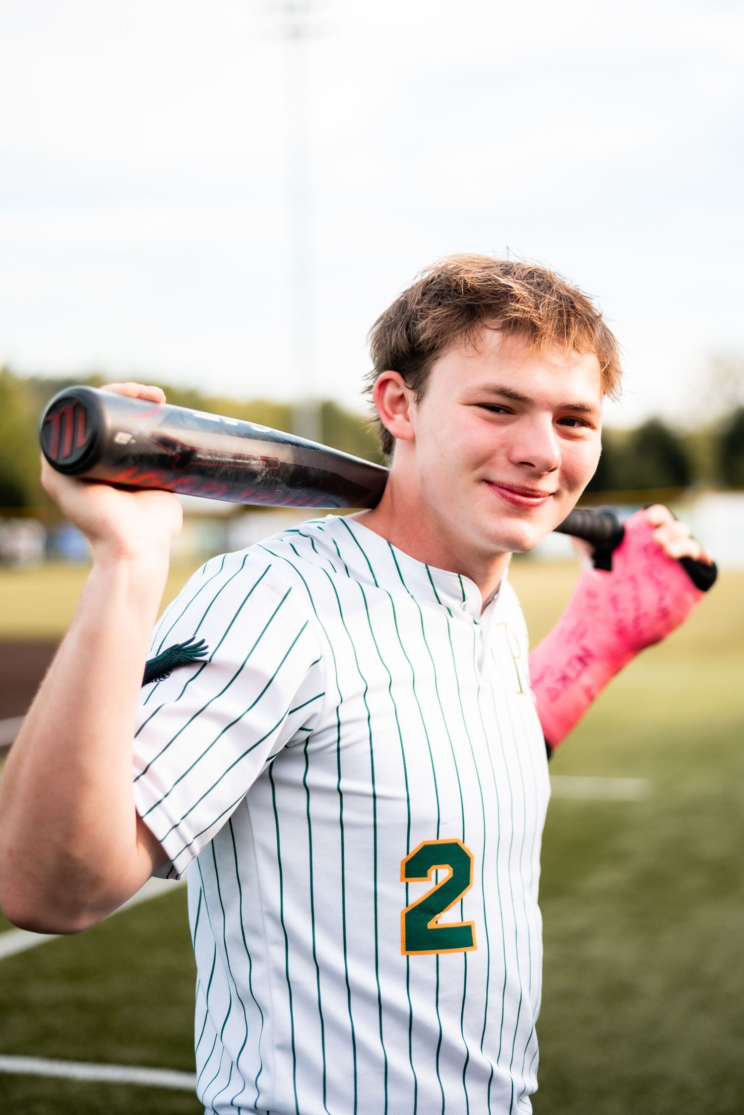 Young baseball player wearing jersey number 2 holds bat over shoulder on field, smiling confidently.