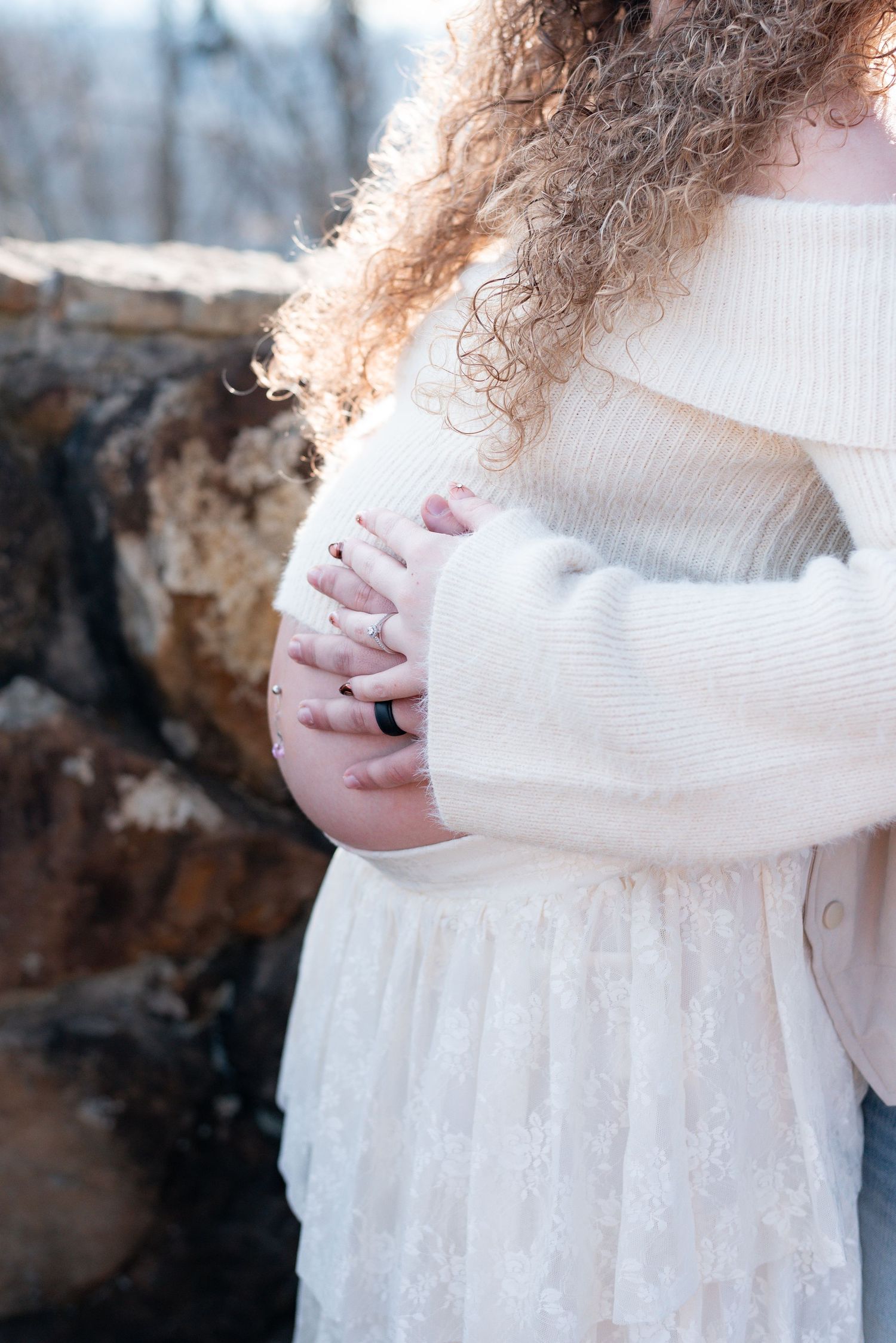 Expectant mother in white sweater dress poses outdoors with natural curly hair.