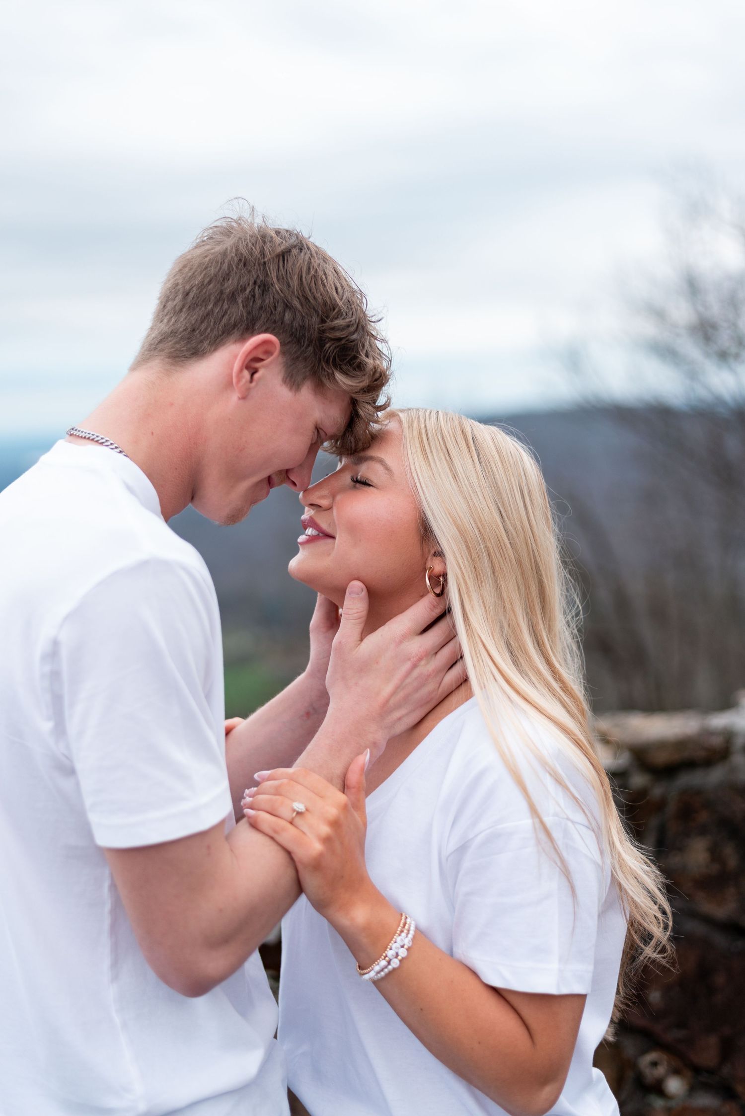 A couple in matching white t-shirts share a romantic moment outdoors, foreheads touching and smiling.