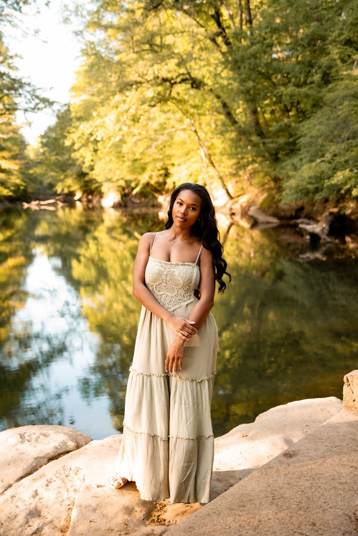 Young woman in cream boho maxi dress standing on rocks by a serene reflective river surrounded by lush green trees.