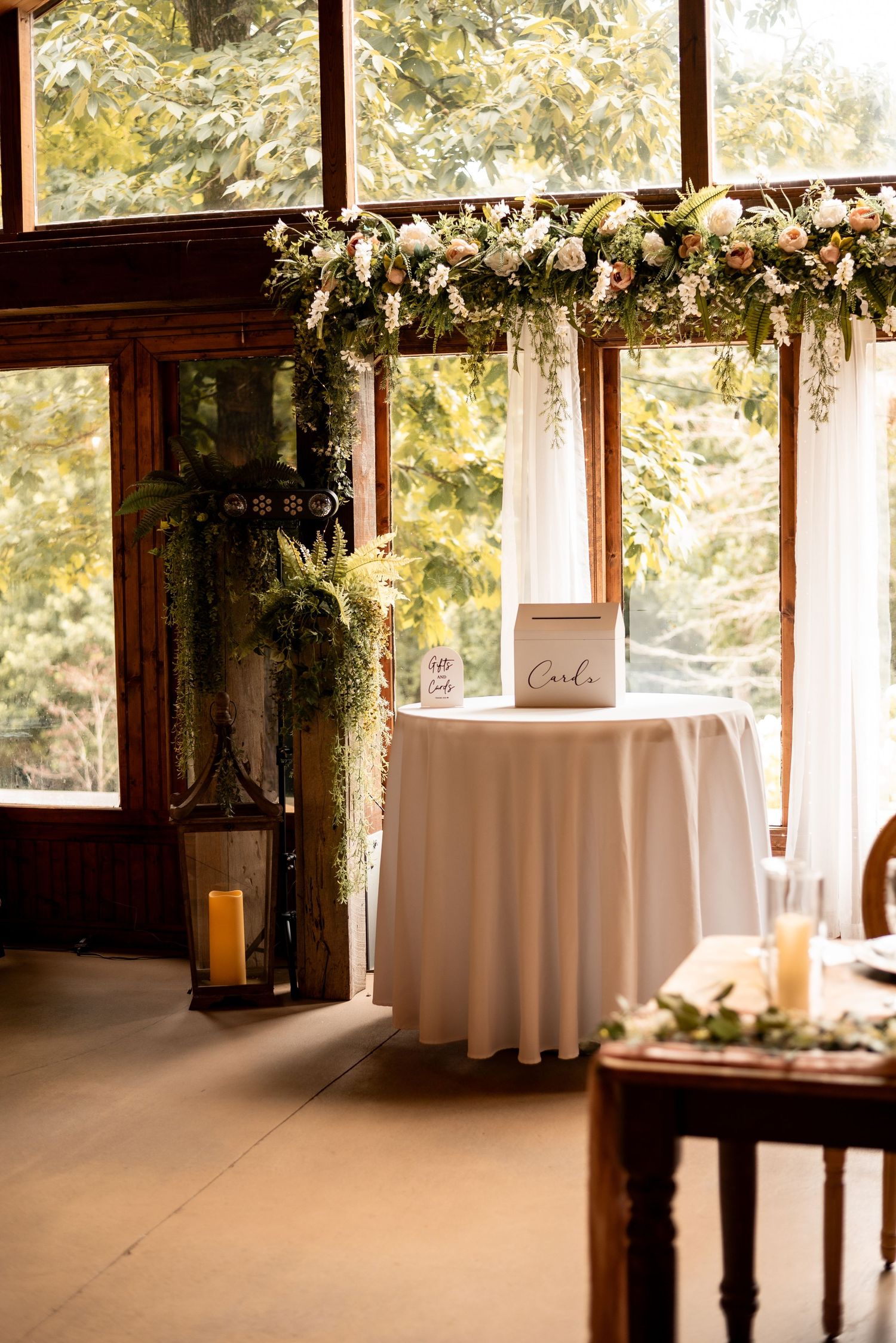 Elegant wedding reception table with white linen, card box, floral arch, and greenery near sunlit windows.