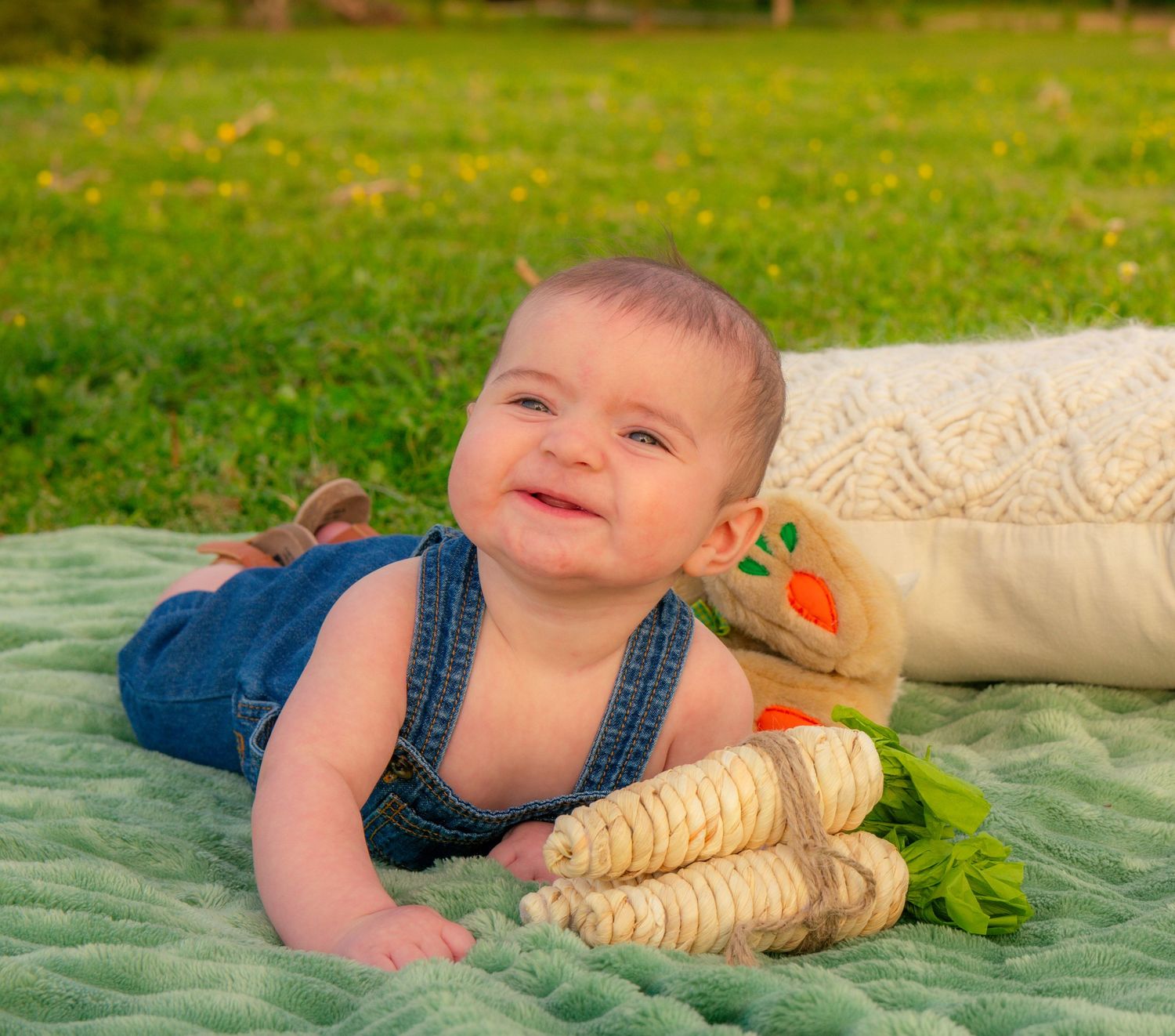Baby lying on tummy on green blanket with plush toy outdoors.