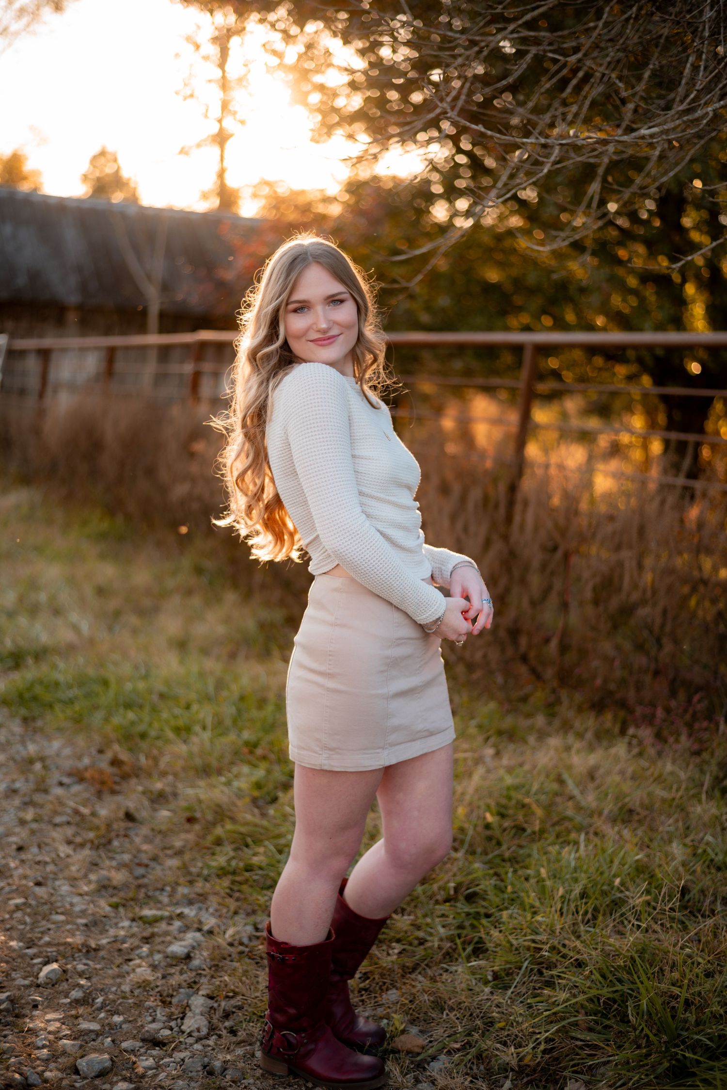 Person in white sweater and boots poses playfully in a field during golden hour.