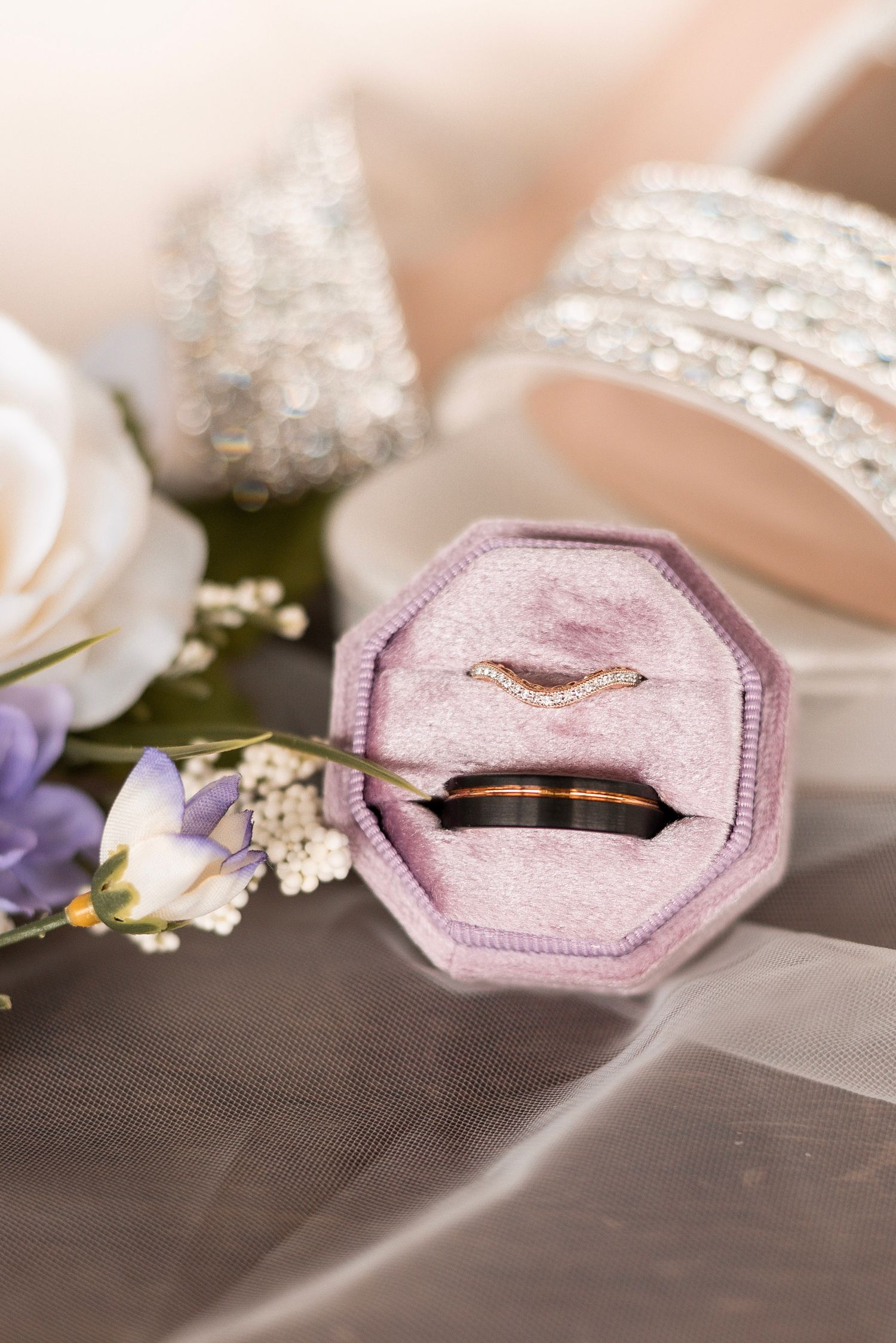 Two wedding rings in an octagonal pink velvet box surrounded by white roses and purple flowers on a bridal veil.