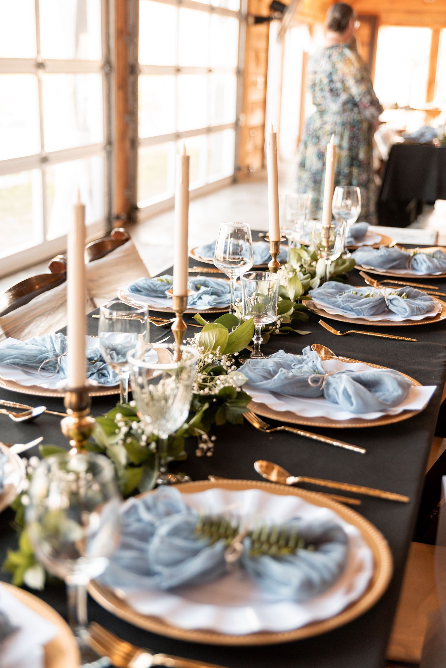 Elegant wedding table setting with blue napkins, gold chargers, greenery runner, and tall candles on black tablecloth.
