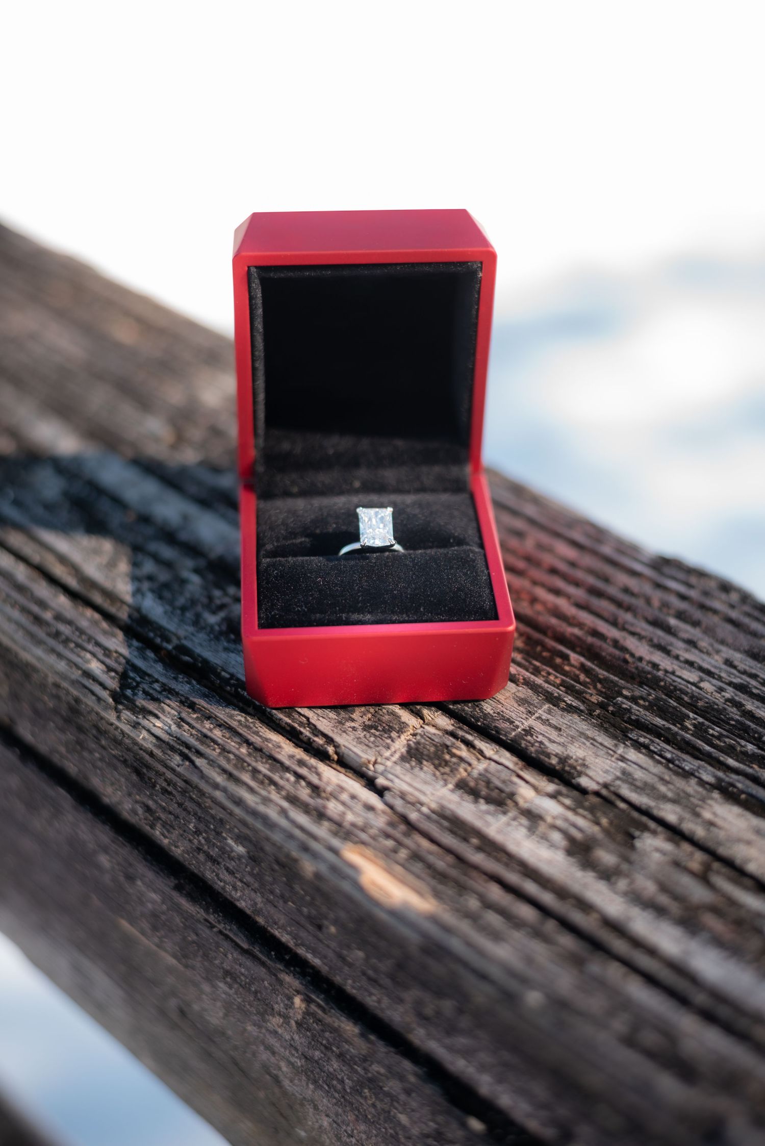 Diamond engagement ring in a red velvet box displayed on weathered wood.