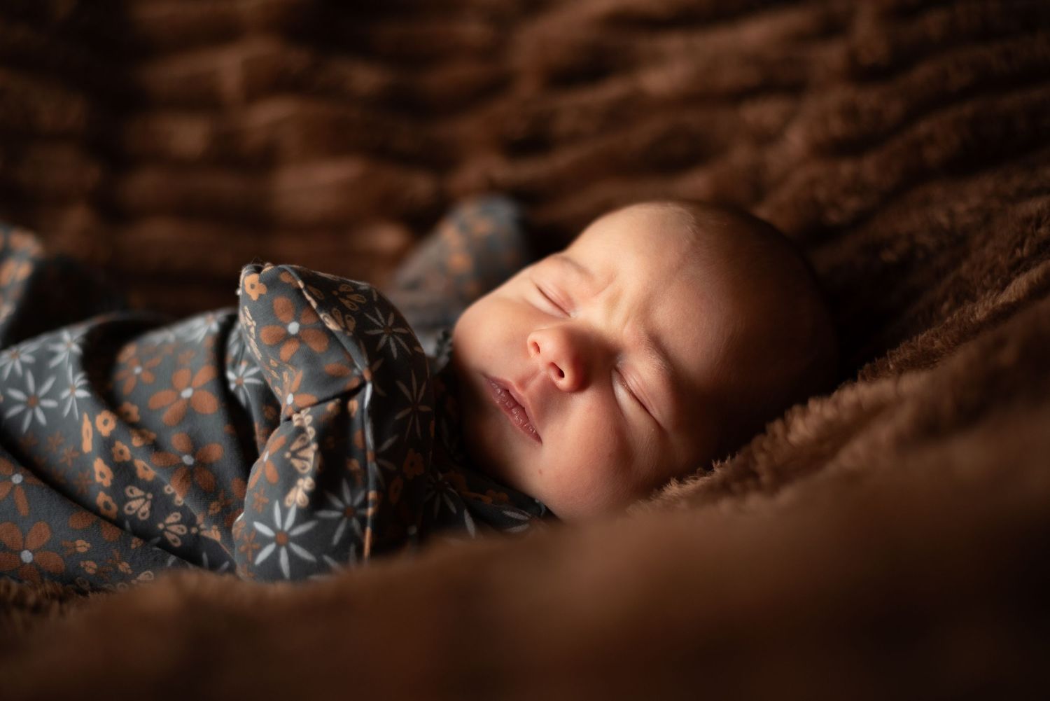 Peaceful sleeping newborn on dark brown textured blanket with dramatic lighting.
