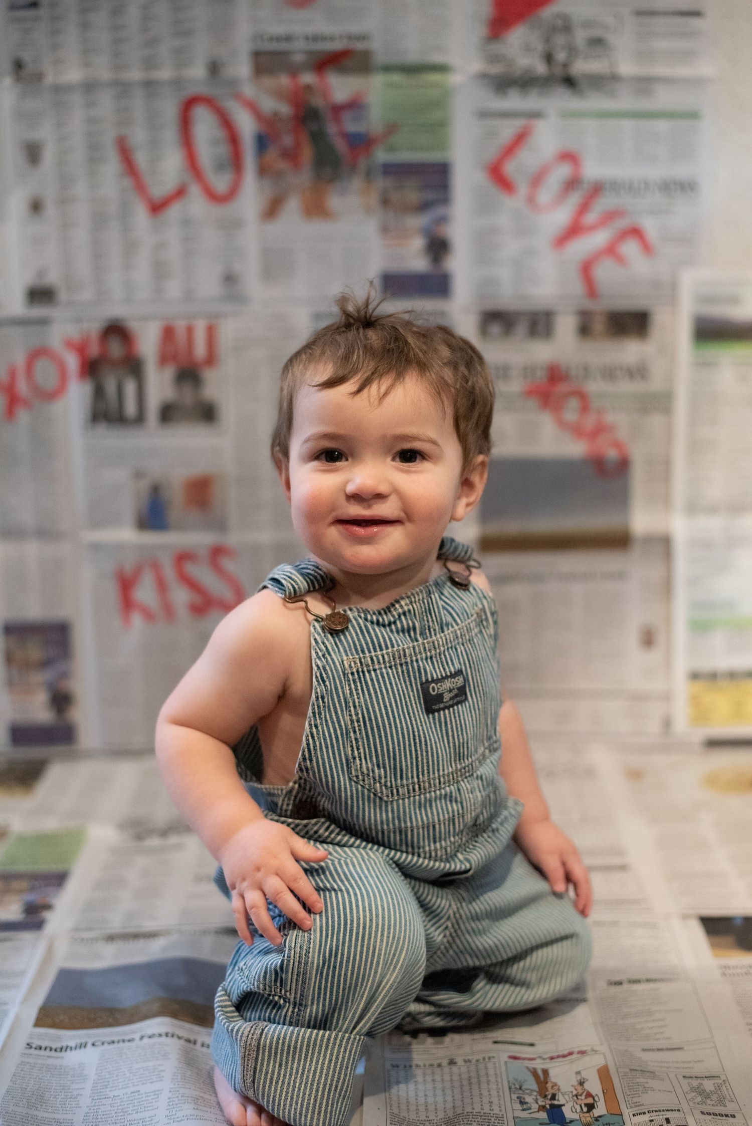 A toddler in striped overalls sits on newspaper-covered floor with love messages on walls.