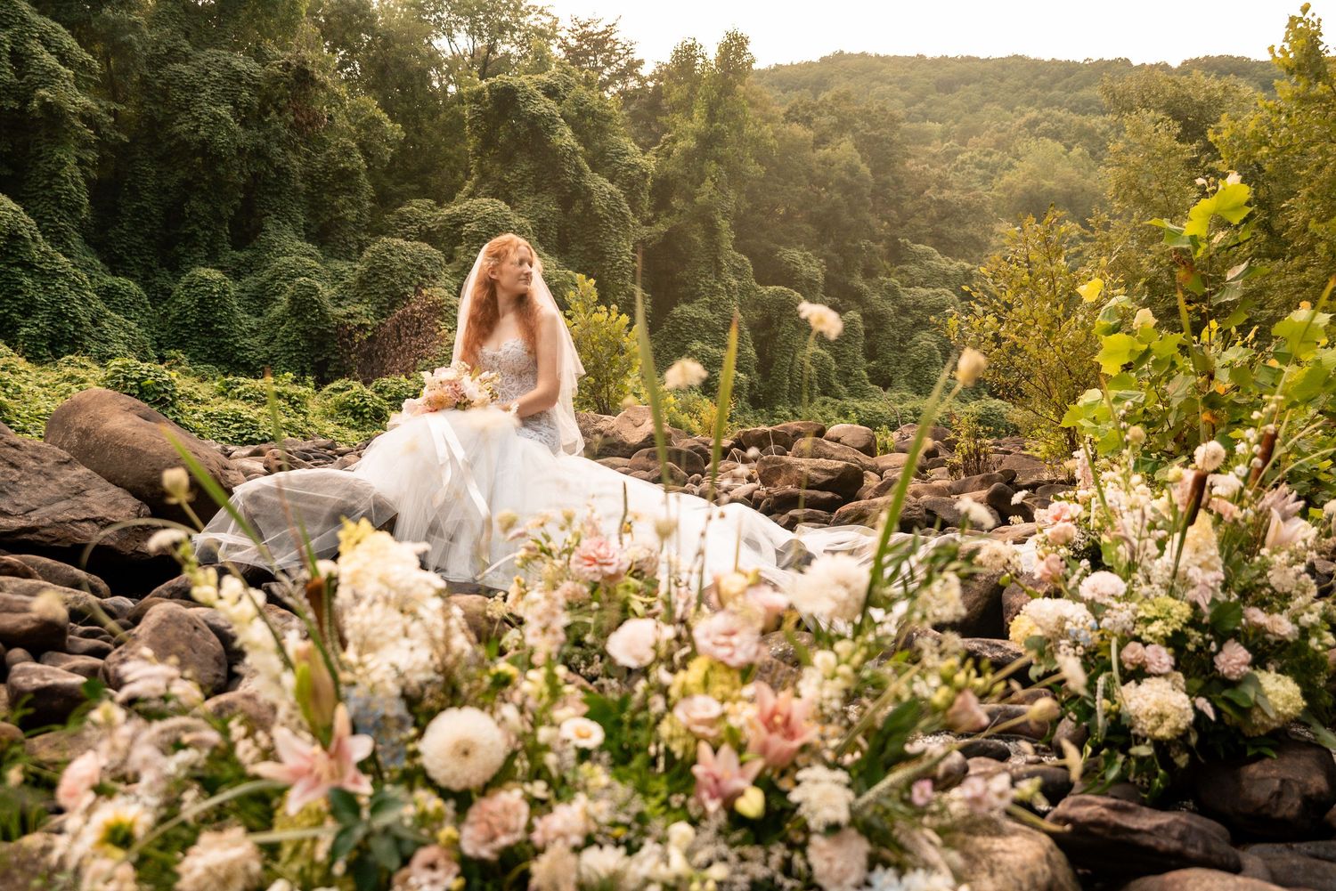 Bride in white gown sits among lush greenery and pink florals on rocky terrain near a waterfall.