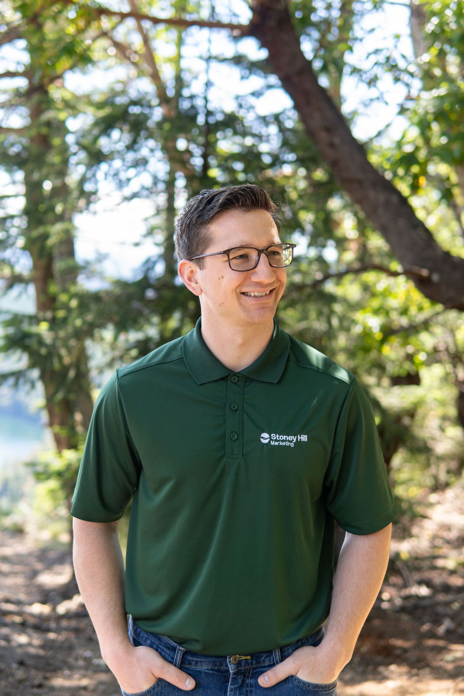 Smiling man in green Stoney Hill polo shirt standing outdoors among trees in a natural setting.