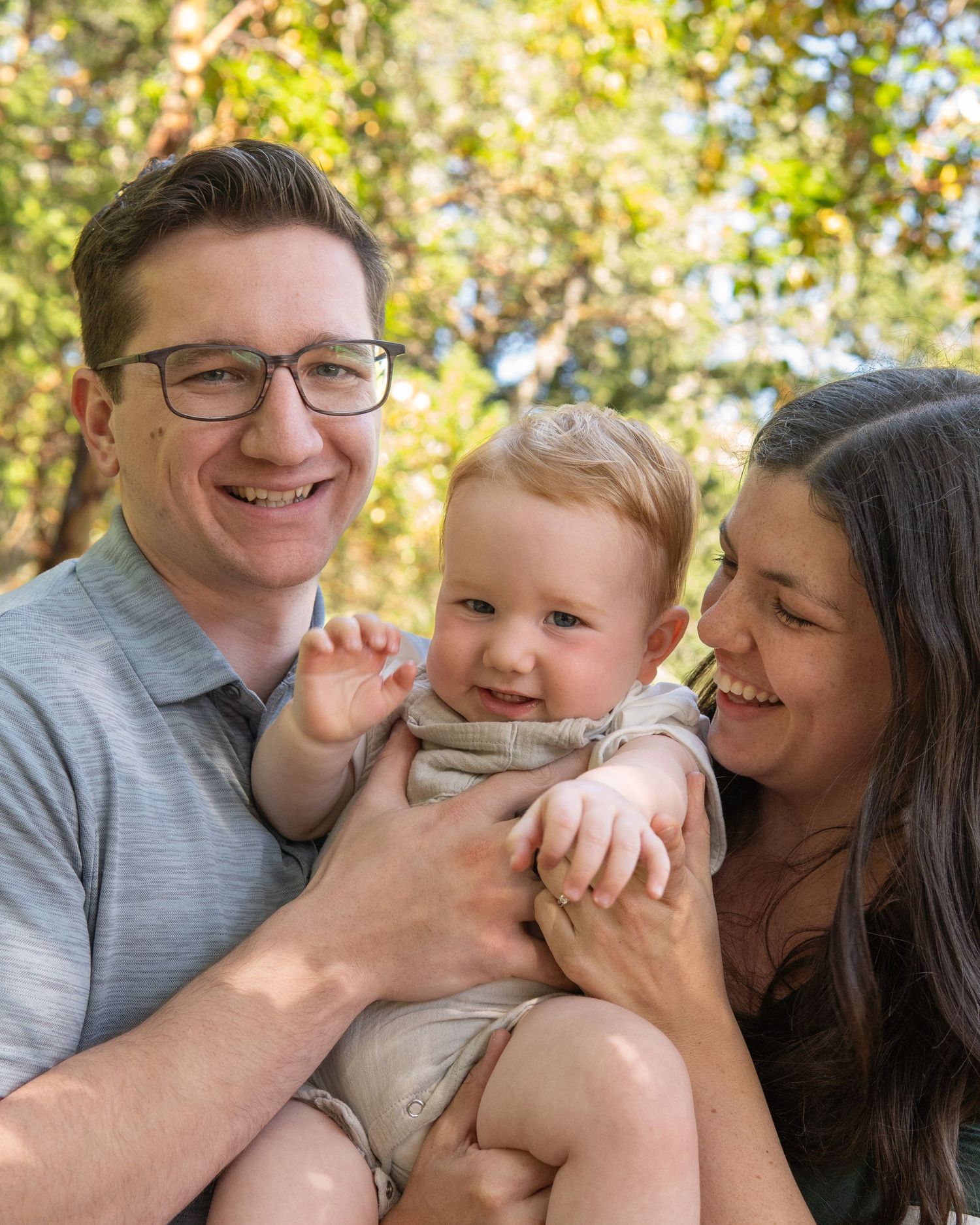 Happy family portrait with smiling parents holding their cheerful baby outdoors among green trees.