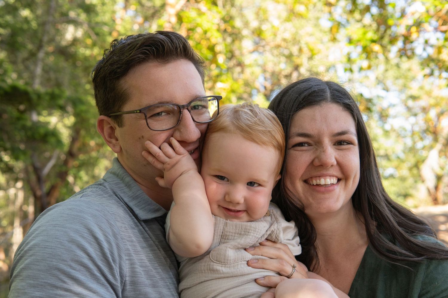 Happy family portrait with smiling parents holding a cute baby outdoors among green trees.