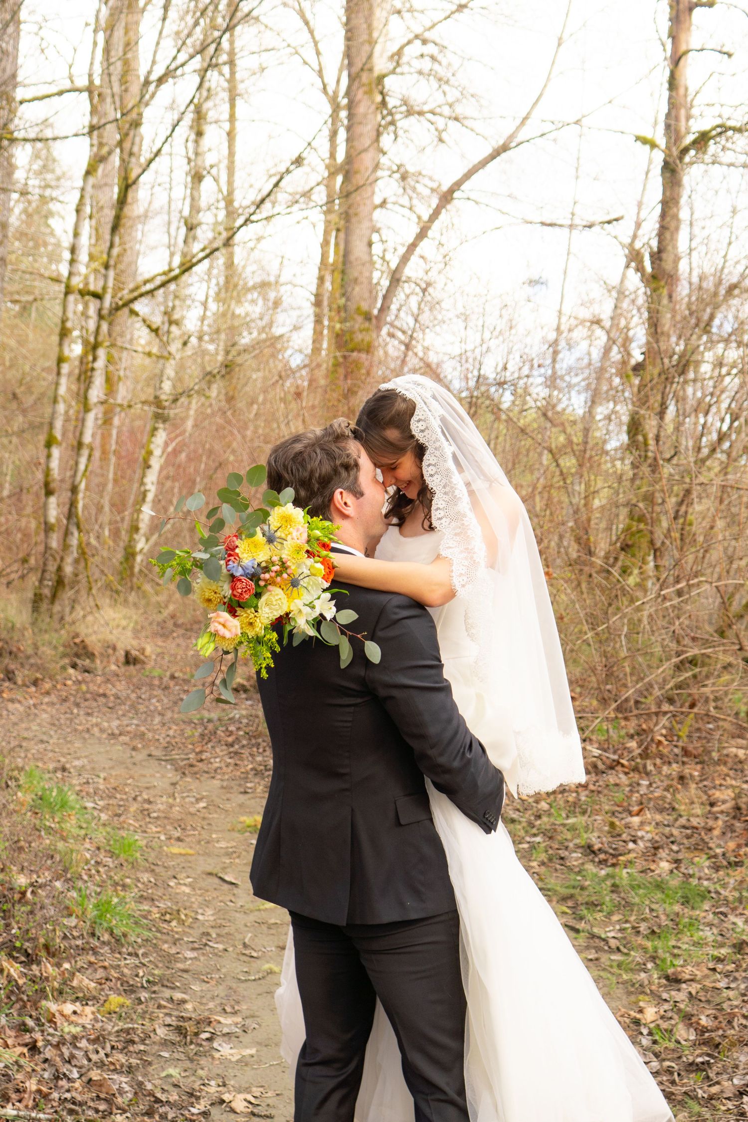 Newlywed couple embracing and kissing on a forest path, bride holding a colorful bouquet.