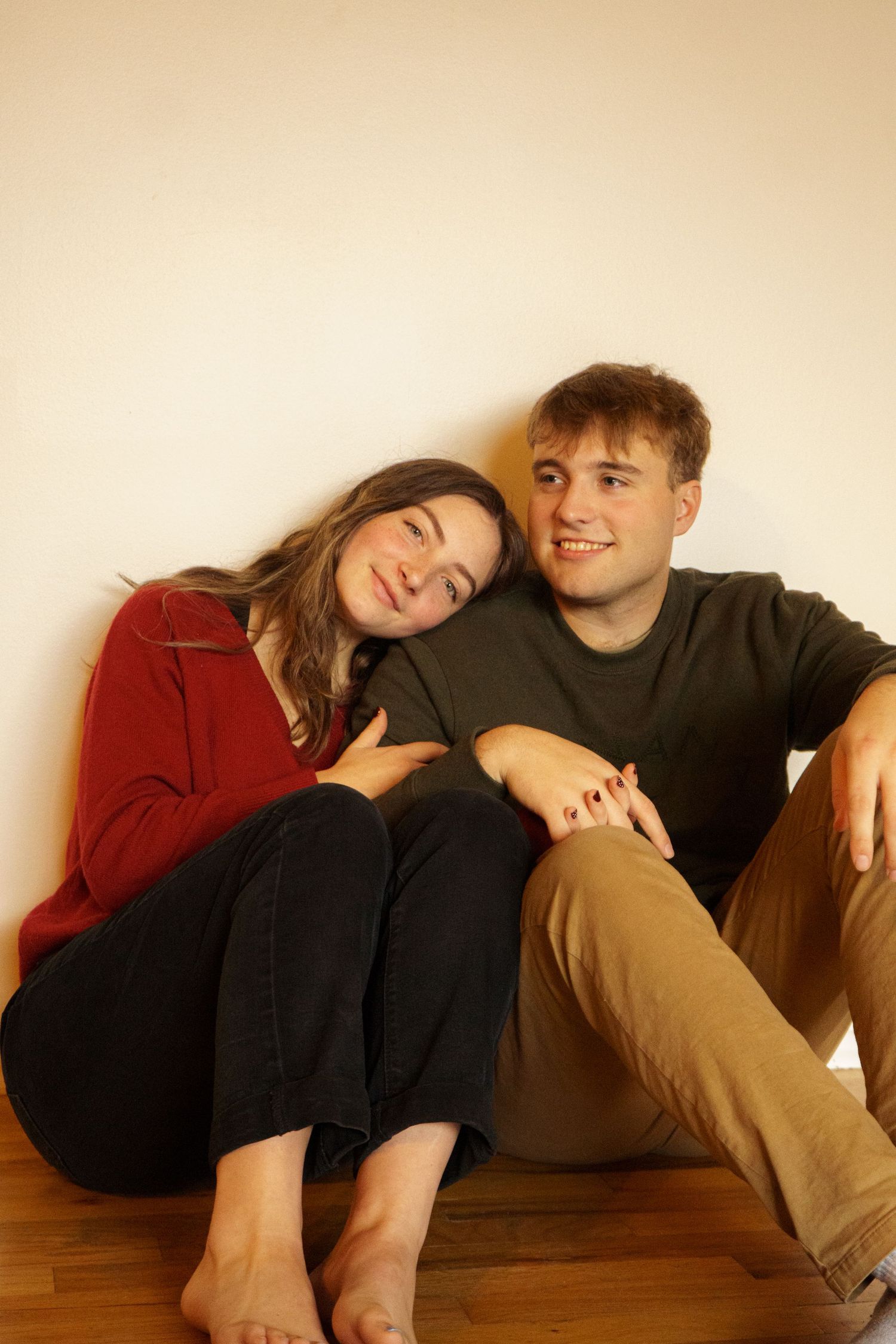 A young couple sits on a hardwood floor, the woman in red leaning on the man in an olive sweater, holding hands.