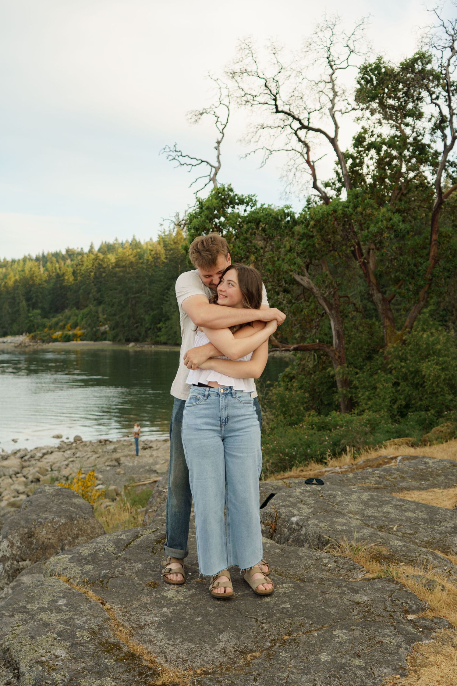 Couple embracing on rocky shore with lush green trees and calm lake in background.